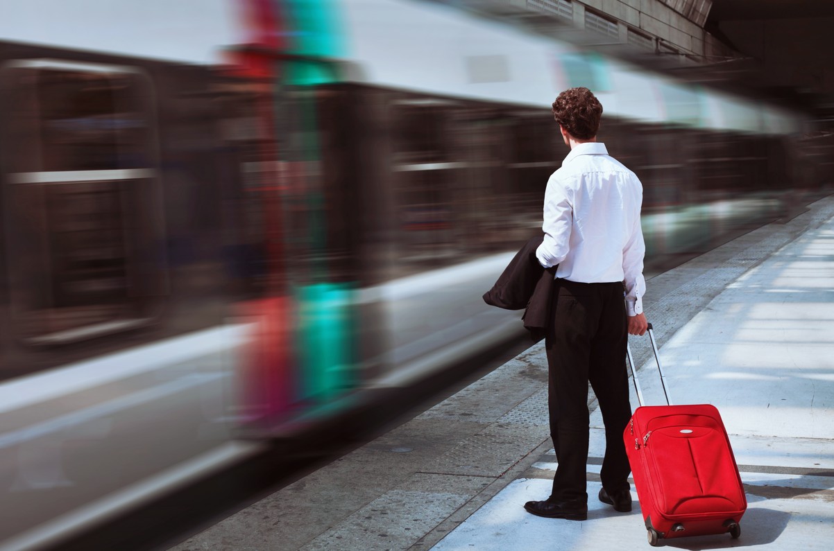A man in a white shirt stands on a New York train platform with a red rolling suitcase as a blur of a train passes by him.