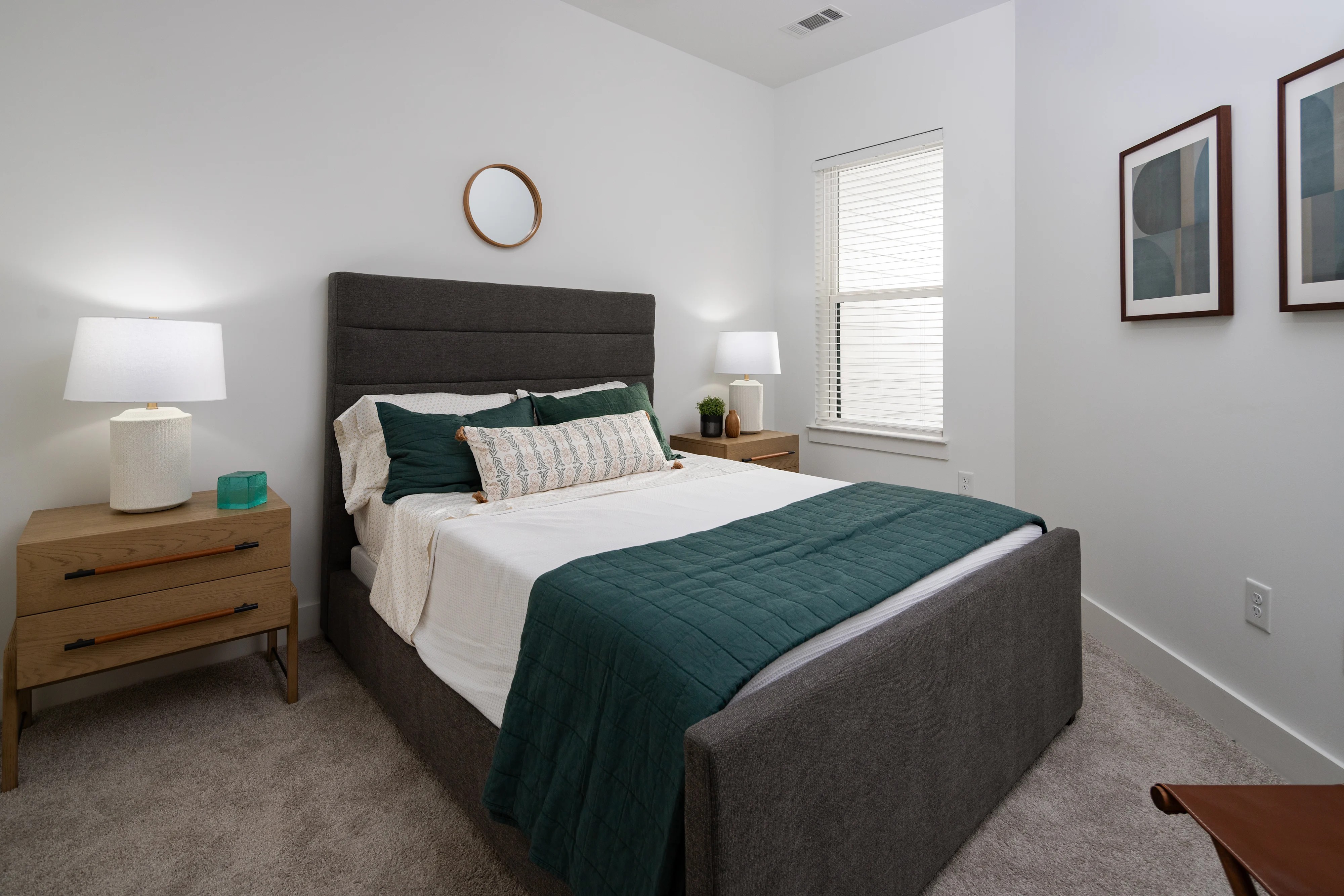 A gray upholstered bed displaying green and white bedding resting between two wood nightstands holding white table lamps.