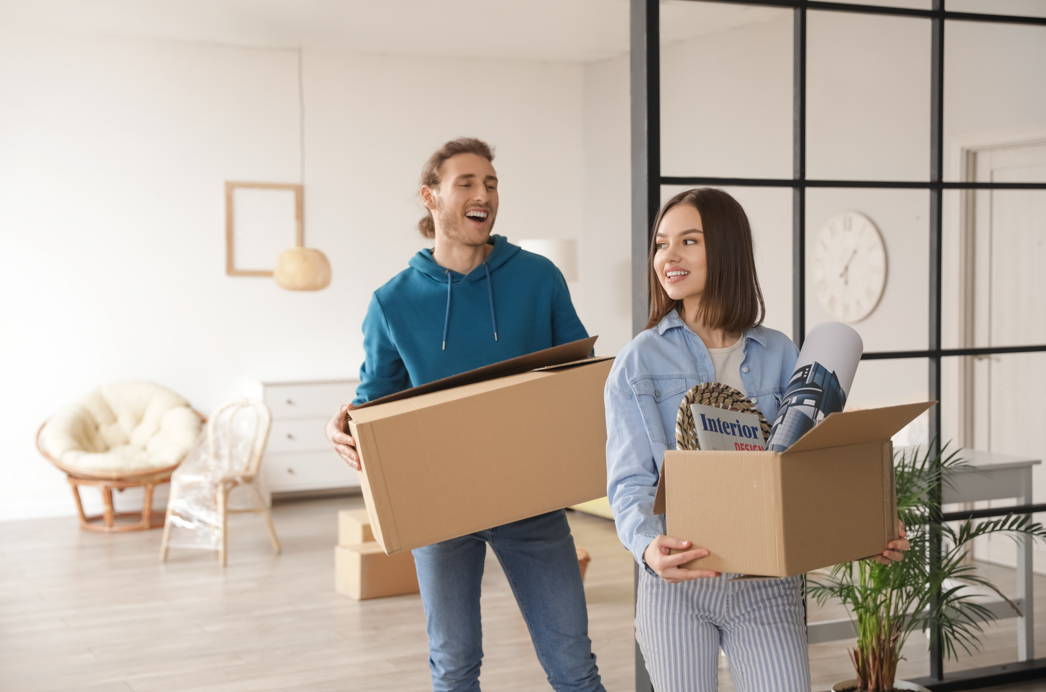 A happy young couple smiling and carrying cardboard moving boxes into their new apartment in New Rochelle, New York.