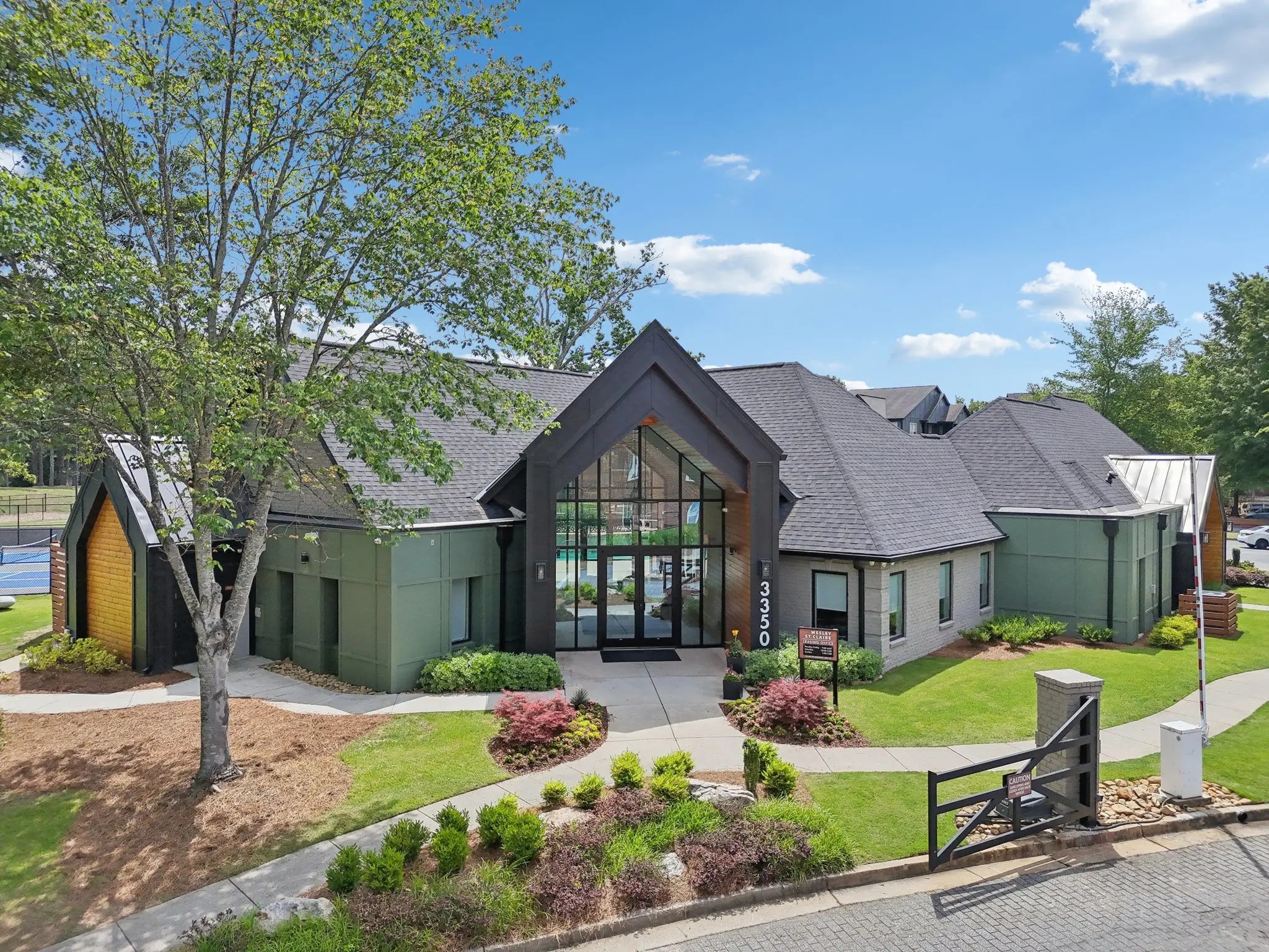 A green and grey clubhouse featuring a dark pitched roof and glass doors resting near green landscaping and trees.