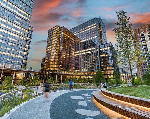 Landscaped pedestrian plaza with a curved walkway and lit bench seating at sunset, framed by glass office towers in National Landing, Arlington VA