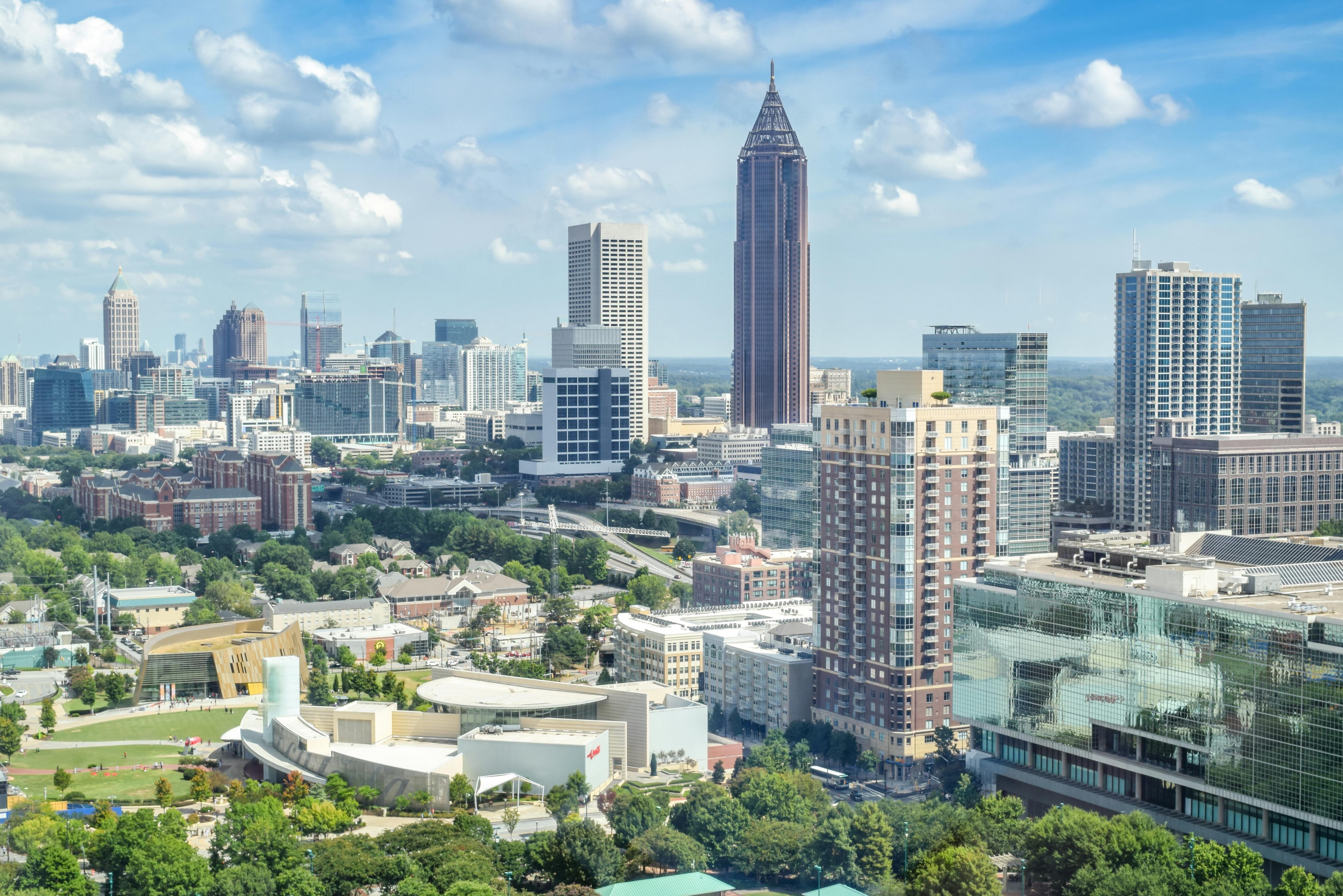 Atlanta skyline featuring tall modern towers, dense greenery, mid-rise buildings, and a bright blue sky with scattered clouds.