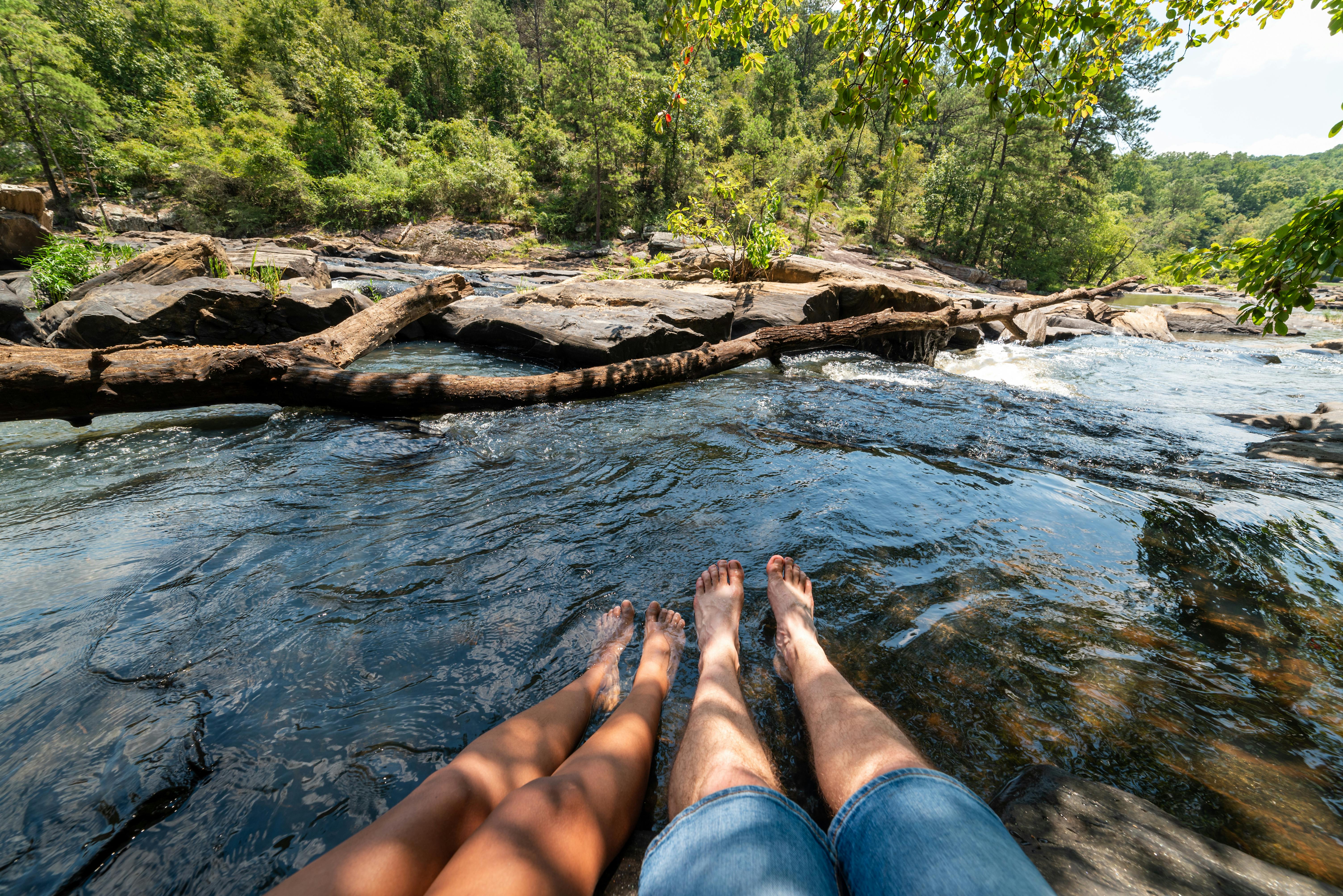 Two pairs of bare legs dangling into a flowing river near a thick fallen log crossing the water in a green forest.
