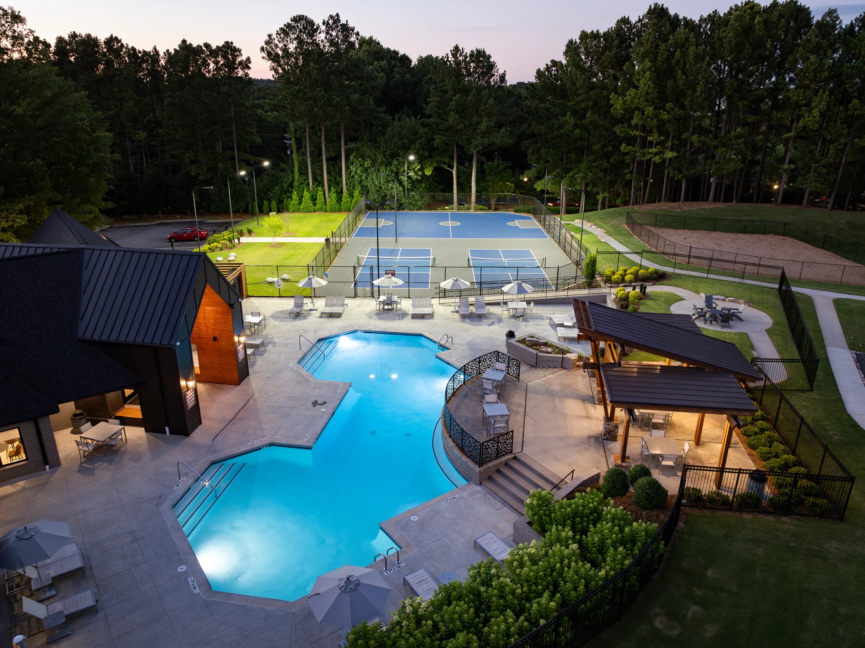The Wesley St Claire clubhouse sitting near a glowing swimming pool and outdoor tennis courts bordering tall green trees.
