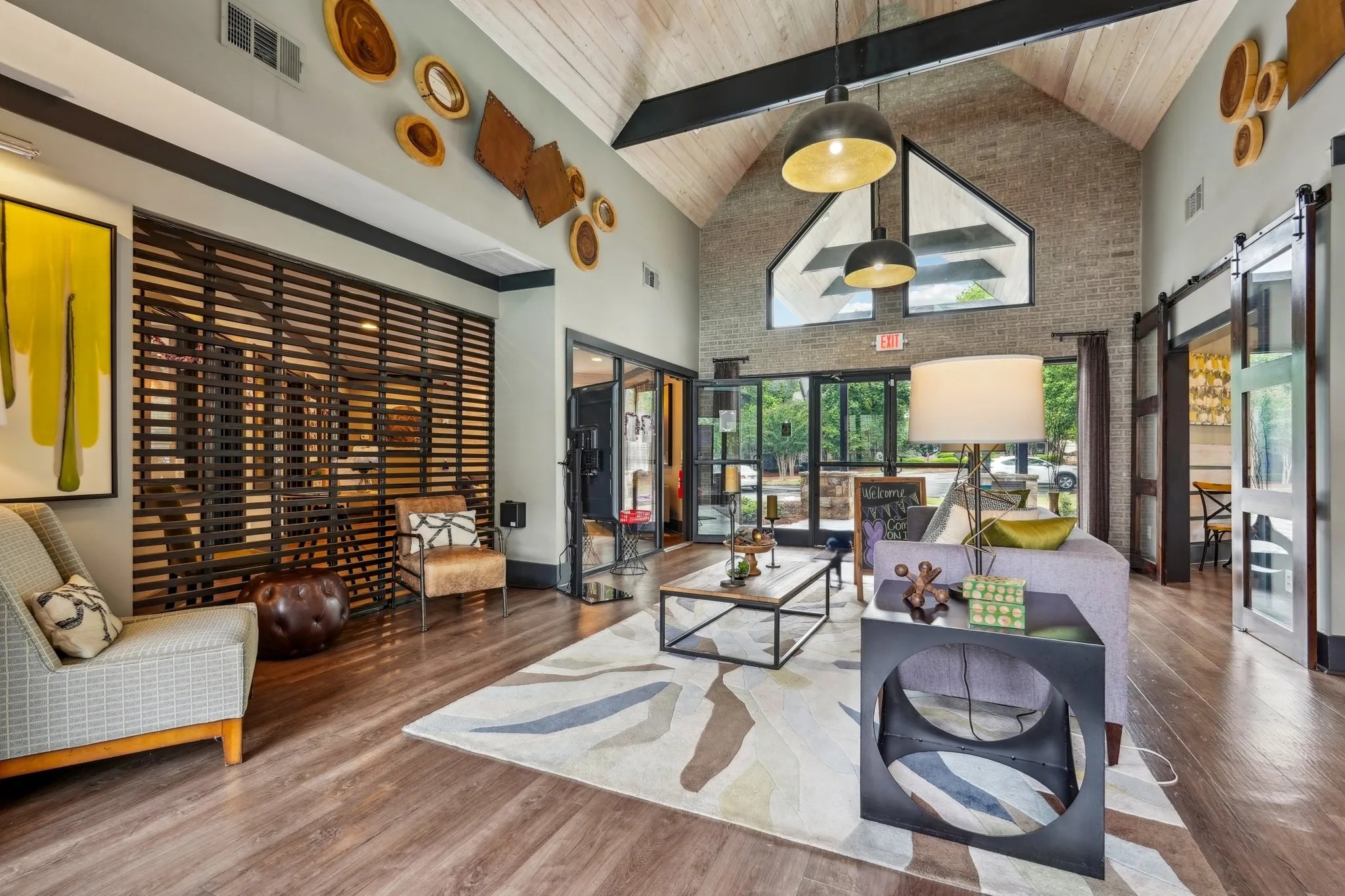 An indoor lounge featuring a high ceiling and modern furniture resting on a patterned rug near large glass doors.