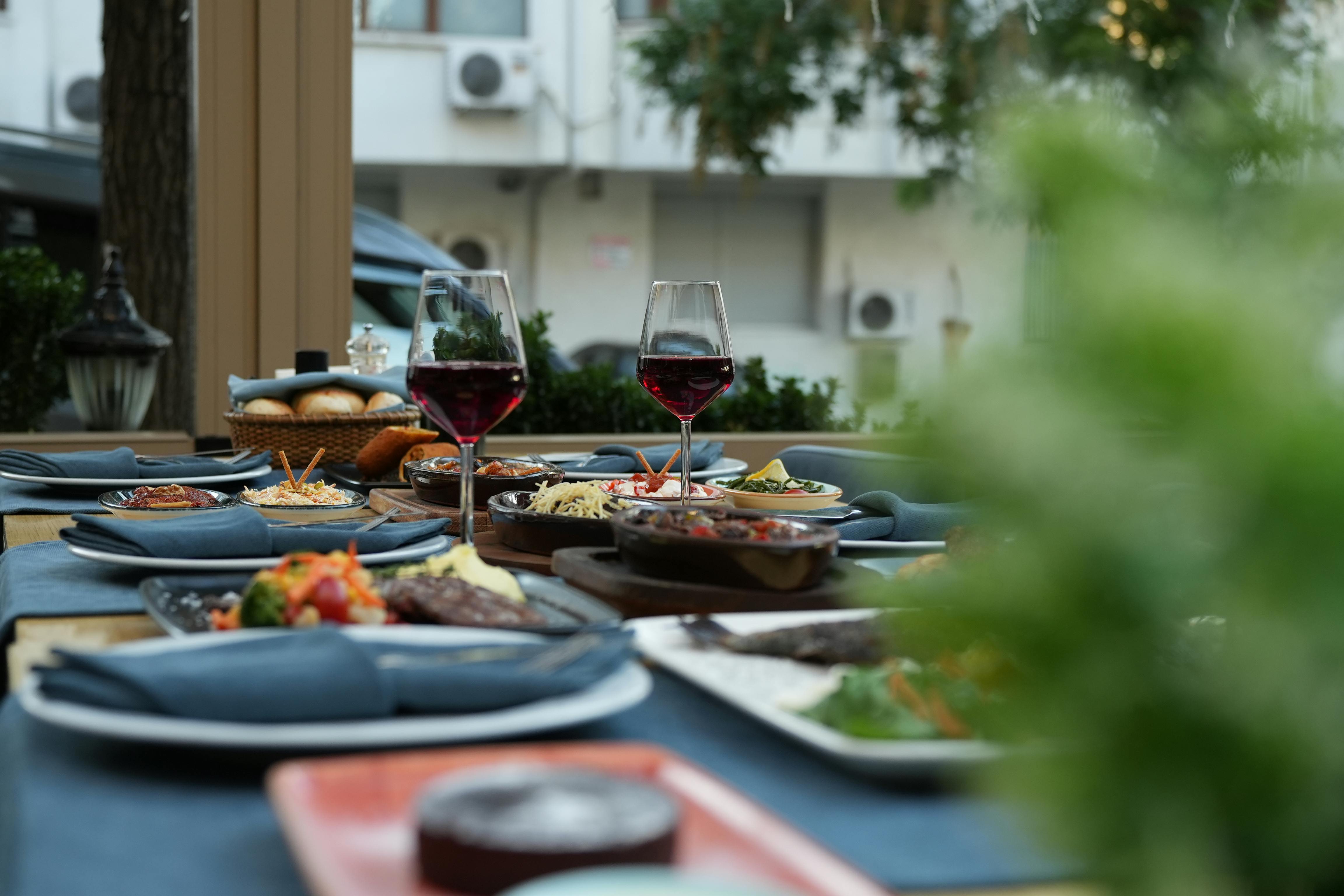 Two glasses of red wine sitting among plates of food and folded blue napkins resting on an outdoor dining table.