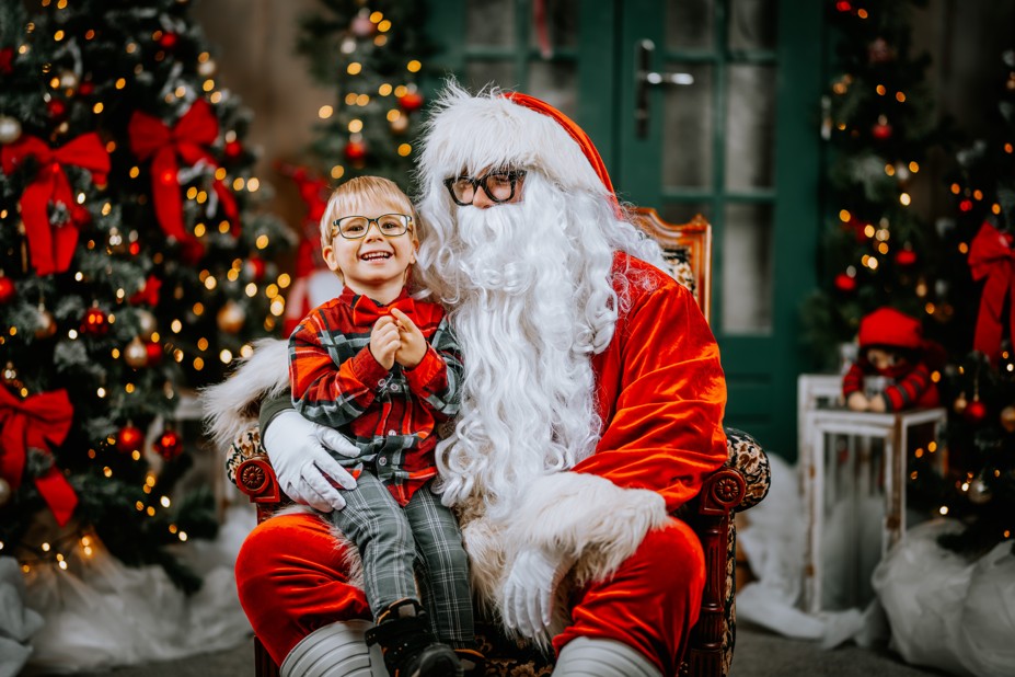 A young child smiling brightly while sitting on Santa Claus's lap in a festive scene decorated with Christmas trees, set in Fairfield, CT.