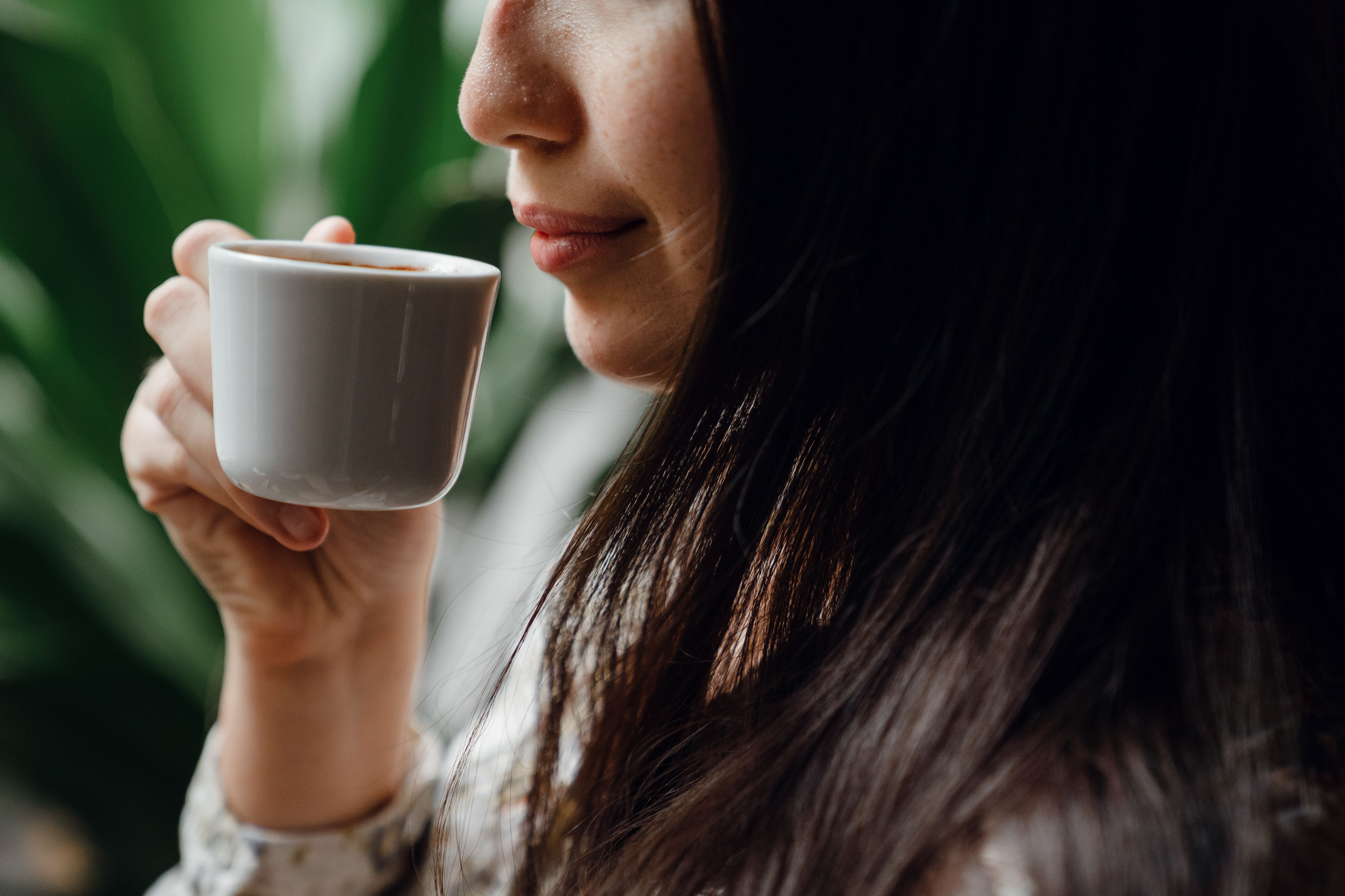 A woman with long dark hair holding a small white ceramic cup while taking a sip of coffee near green plants.