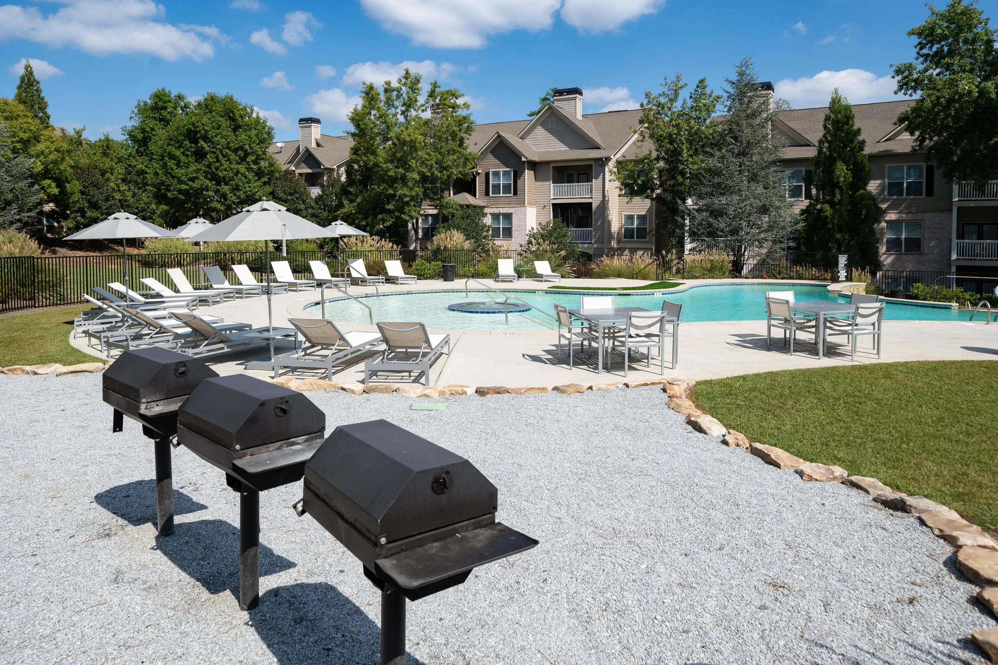 Three black metal grills standing in a gravel area next to a bright blue swimming pool surrounded by white lounge chairs.