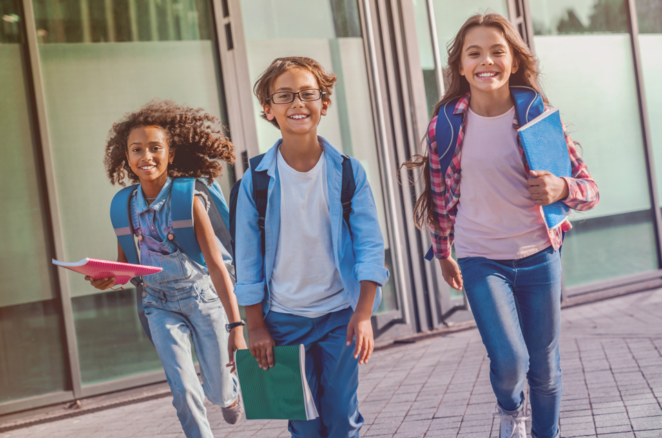 Three smiling students running with backpacks and notebooks outside school building