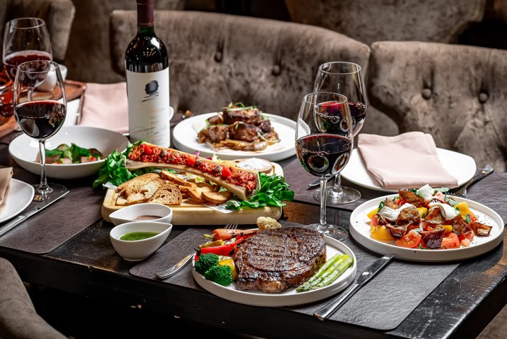 A high-end dinner spread at a restaurant in Asheville, NC, featuring a grilled ribeye steak with asparagus, bone marrow with crostini, various side salads, and glasses of red wine surrounding a bottle of Opus One.