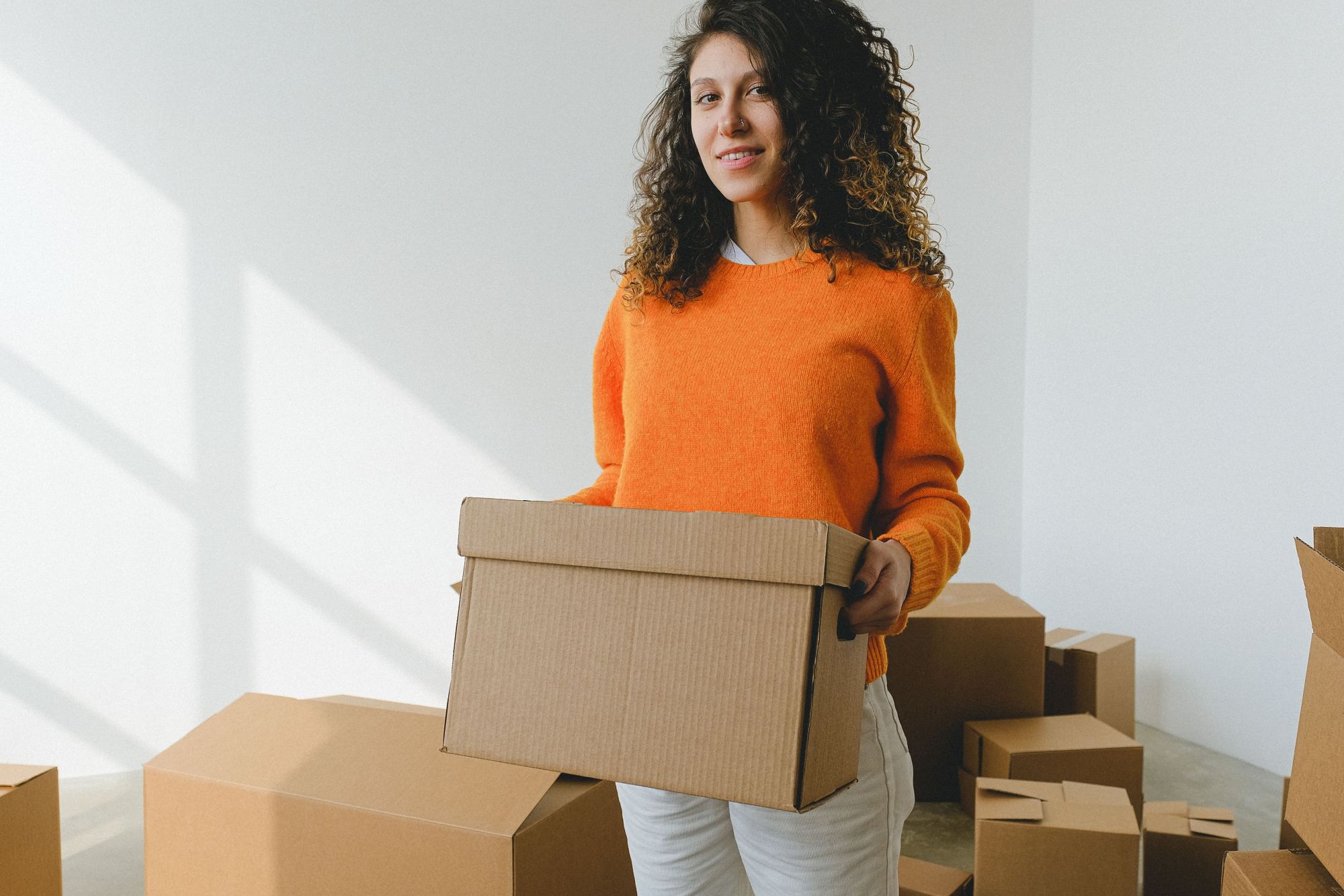 A curly-haired woman wearing an orange sweater holding a closed cardboard box in a white room containing several other boxes.