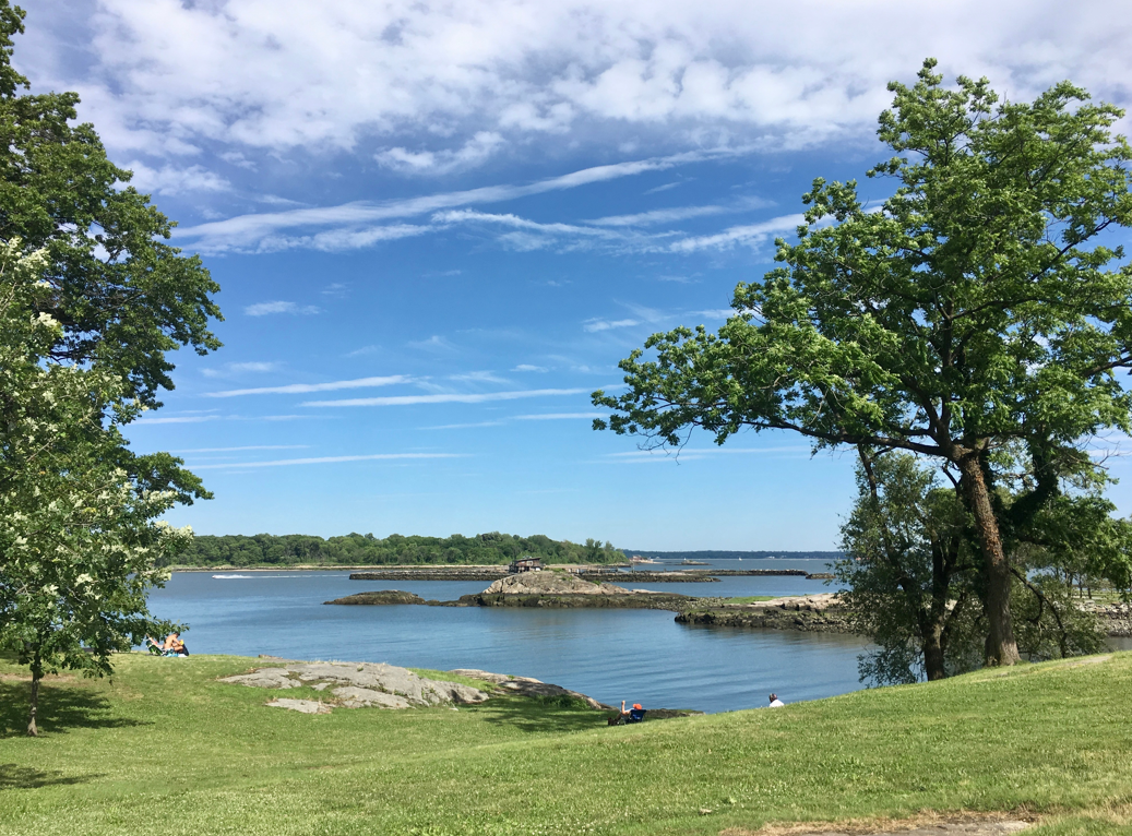 Scenic waterfront view at Glen Island Park in New Rochelle with grassy shoreline, trees, blue sky, and calm Long Island Sound waters
