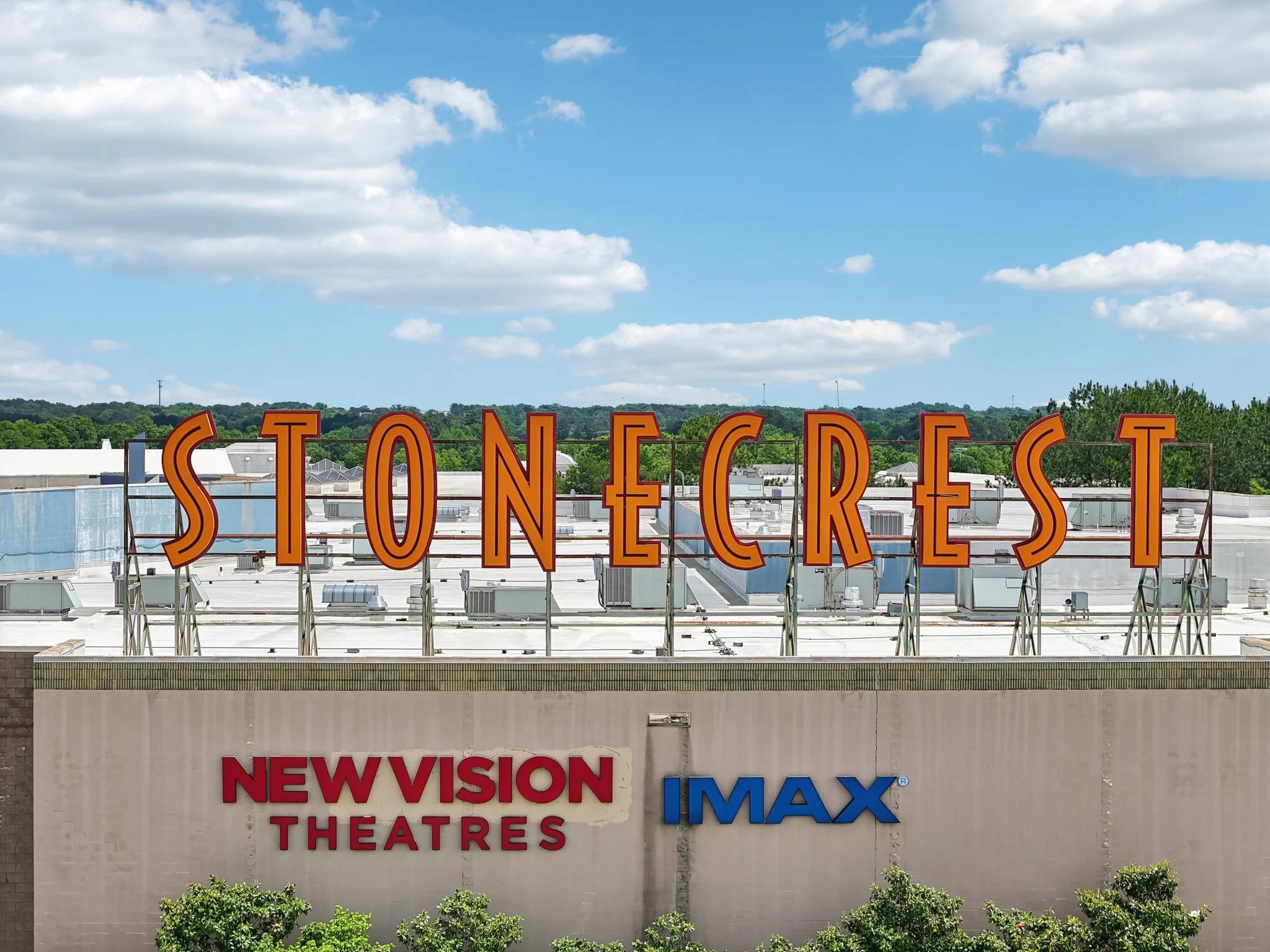 Large orange letters spelling Stonecrest resting on a metal frame above a theater building under a blue cloudy sky.