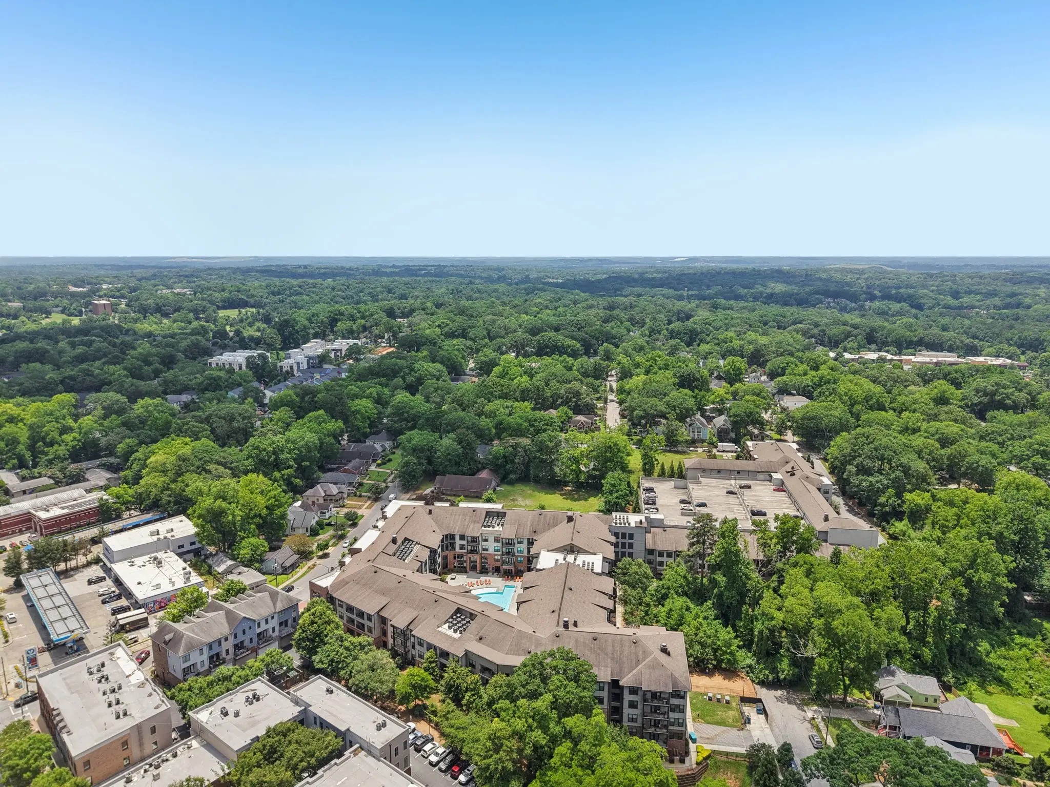 A large apartment complex surrounding a blue swimming pool sitting among dense green trees under a clear sky.