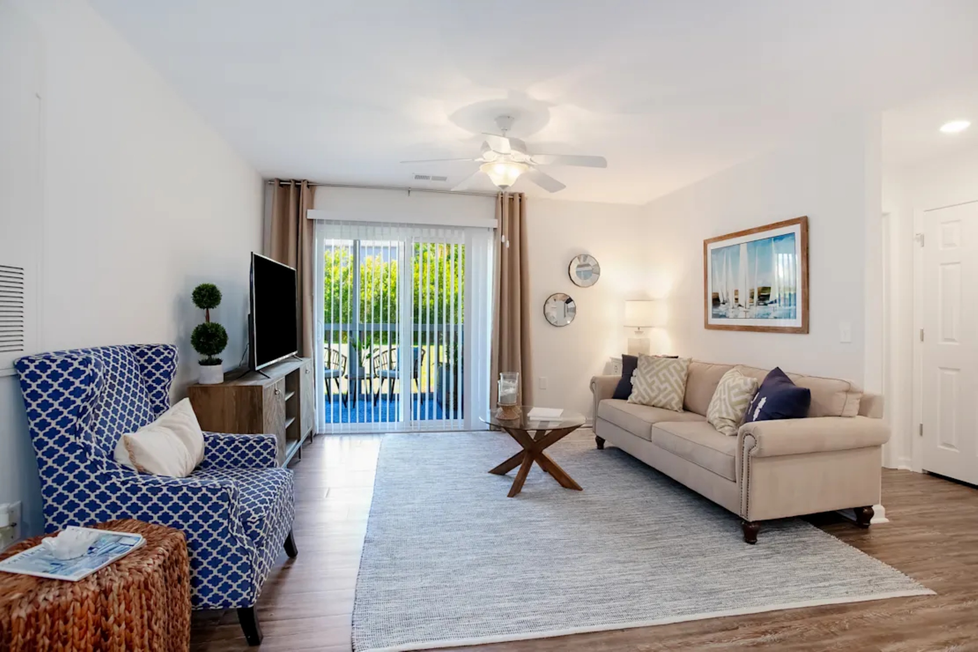 A bright living room featuring a beige sofa, blue patterned armchair, glass coffee table, and sliding glass patio doors.