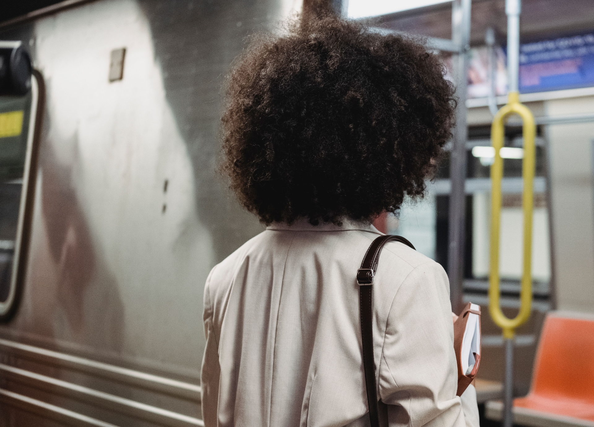 A back view showing a woman with afro hair carrying a shoulder bag while standing outside a public transit train.