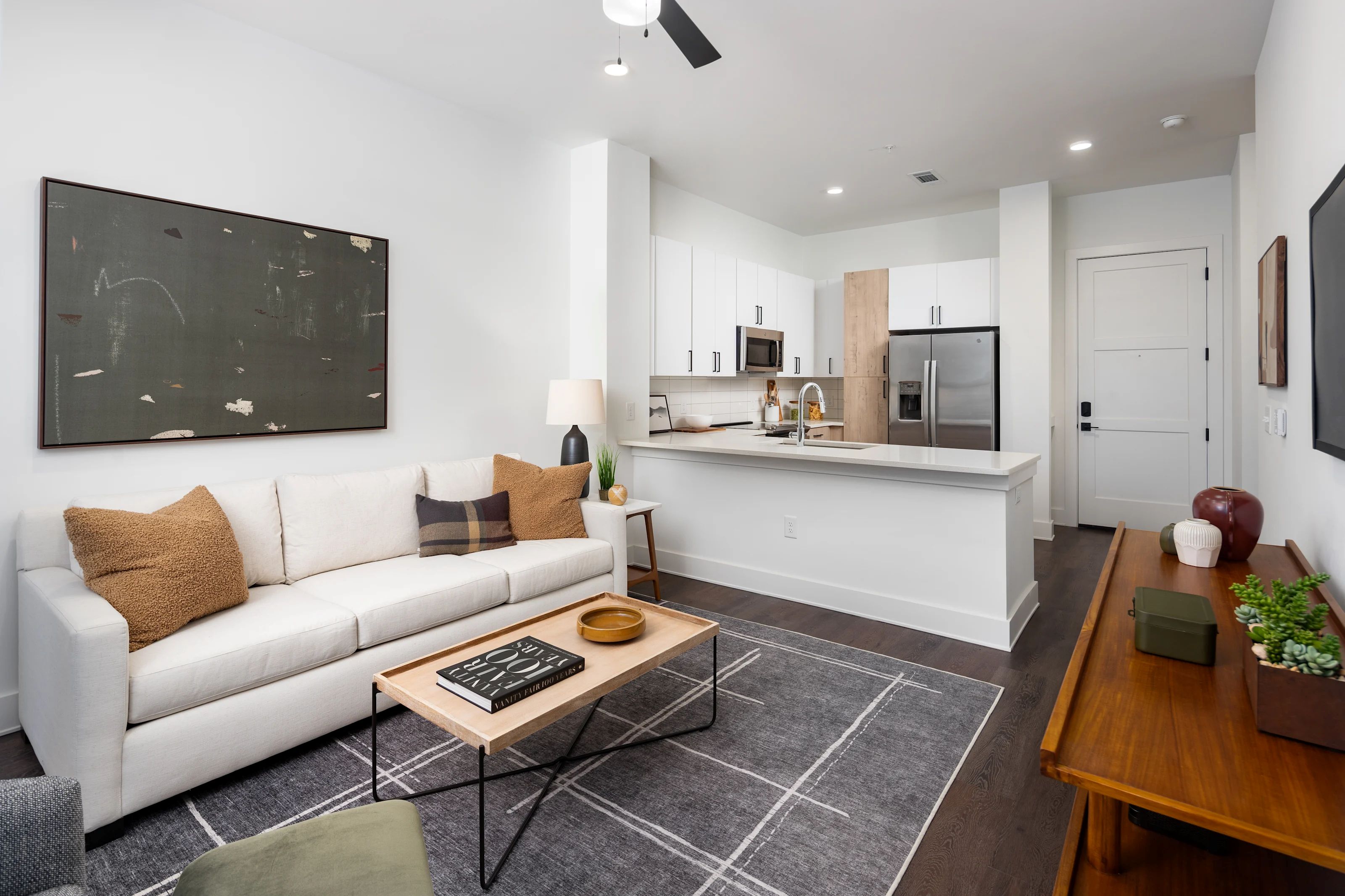 An open living room featuring a white sofa, a wooden coffee table on a patterned rug, and a modern kitchen behind.
