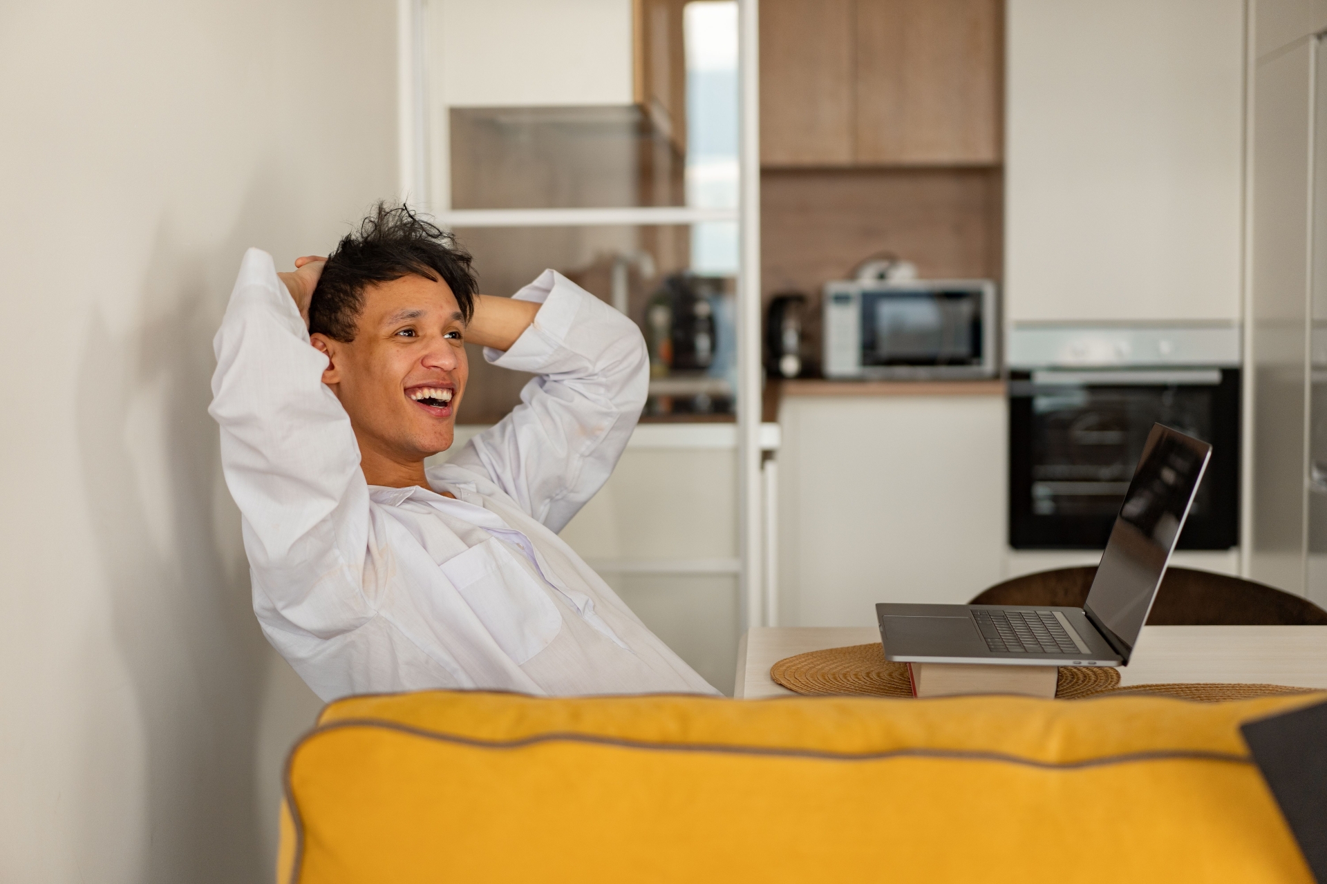 A man sitting at a table with his hands behind his head looking toward an open laptop in front of a kitchen background