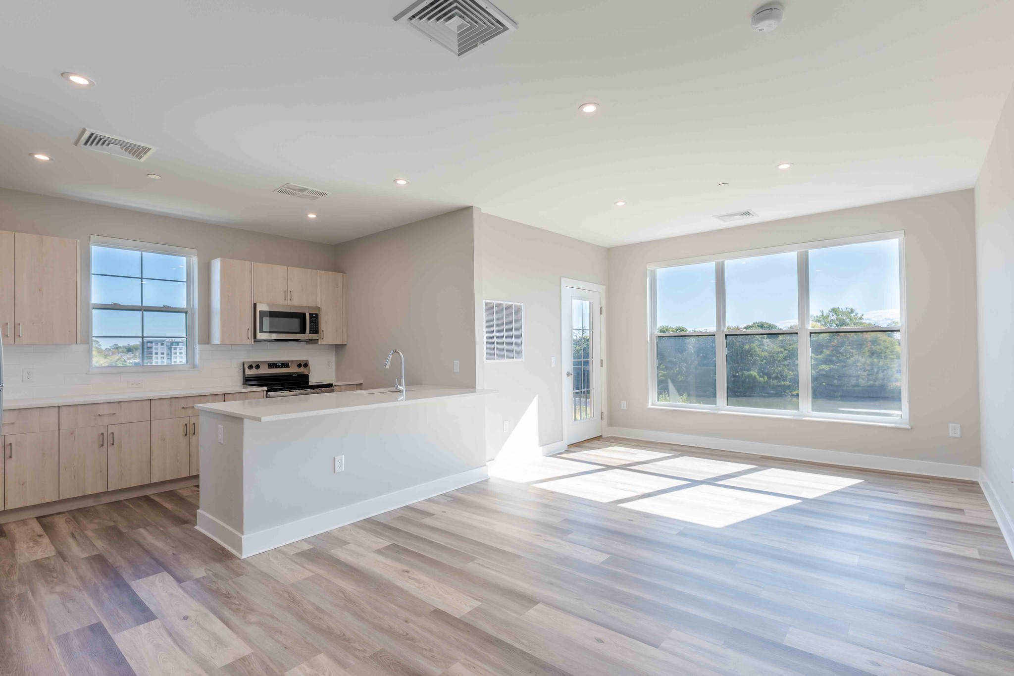 Open-concept kitchen and living area in a unit at Creekline Apartments with light wood flooring, white counters, and large windows.