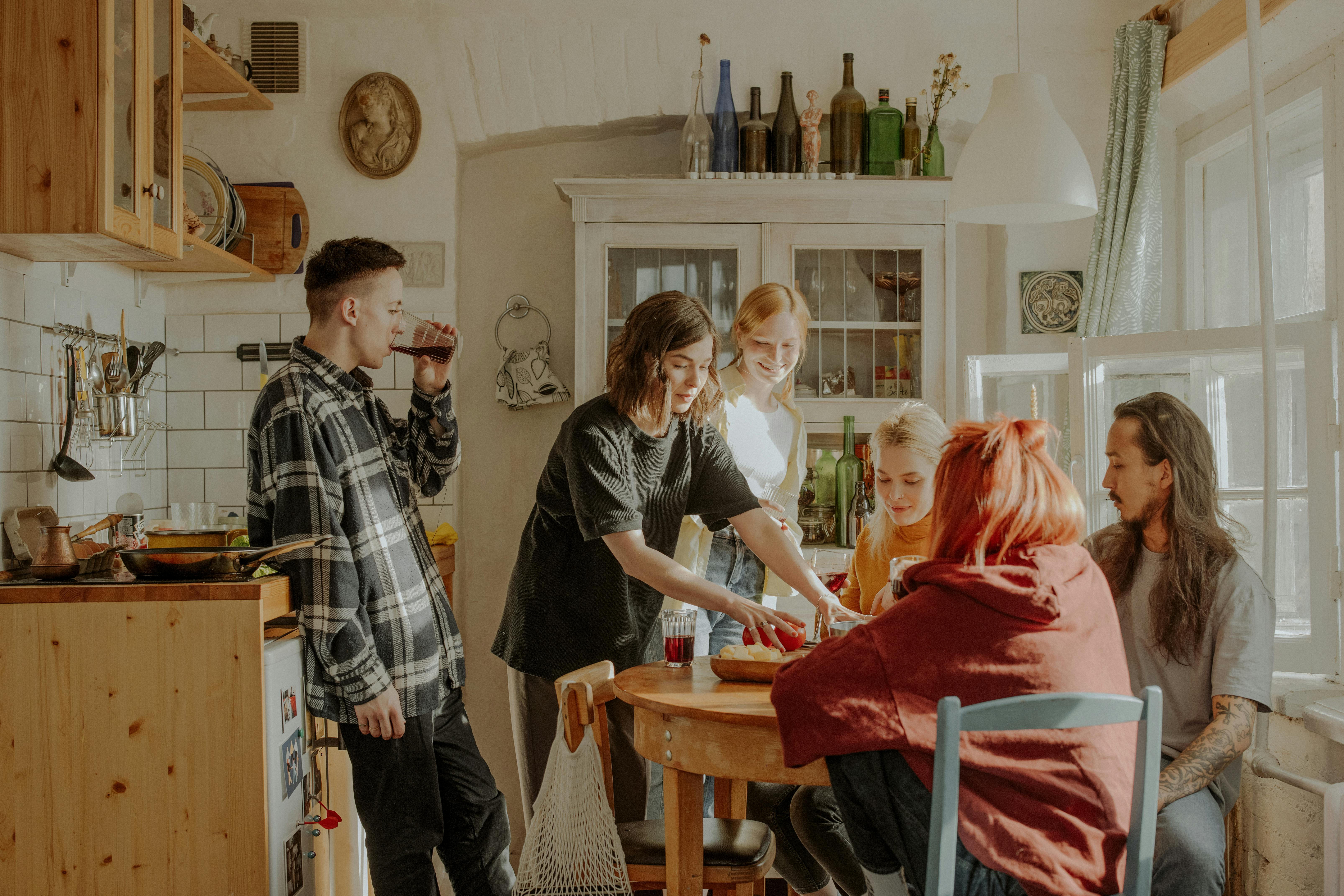 A group of friends wearing casual clothing standing and sitting around a wooden dining table inside a rustic kitchen.