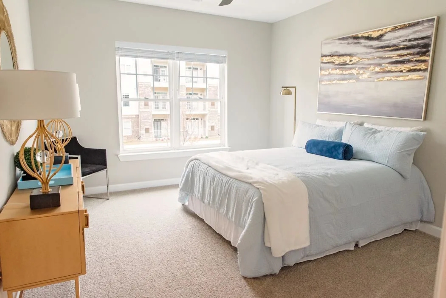 A bright apartment bedroom featuring soft blue bedding, large wall art above the bed, carpet, and stylish lighting.