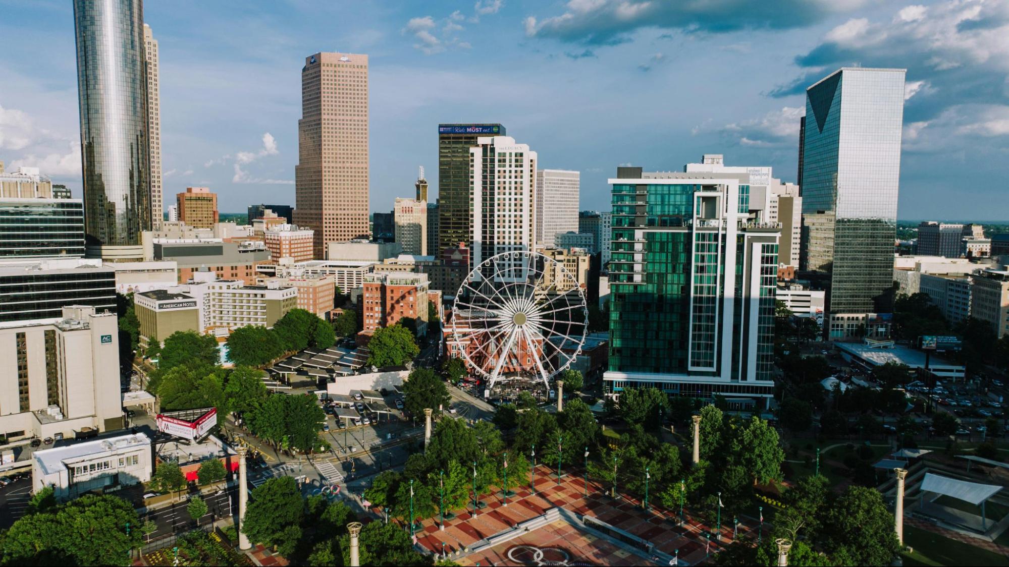 A large white Ferris wheel spinning near tall glass skyscrapers and green trees growing in a sprawling city park.