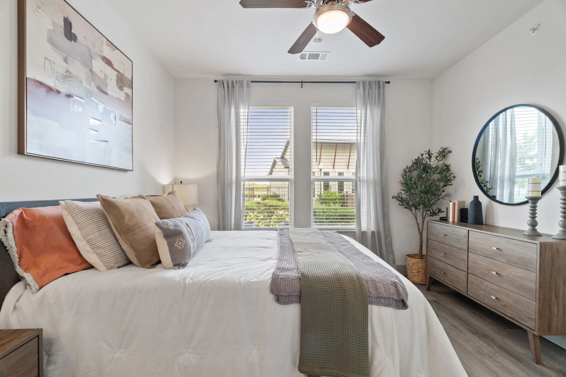A bed featuring colorful pillows resting near a wooden dresser holding a large round mirror and a green plant.