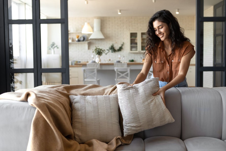 Smiling woman fluffing tan throw pillows on gray sofa in bright living room