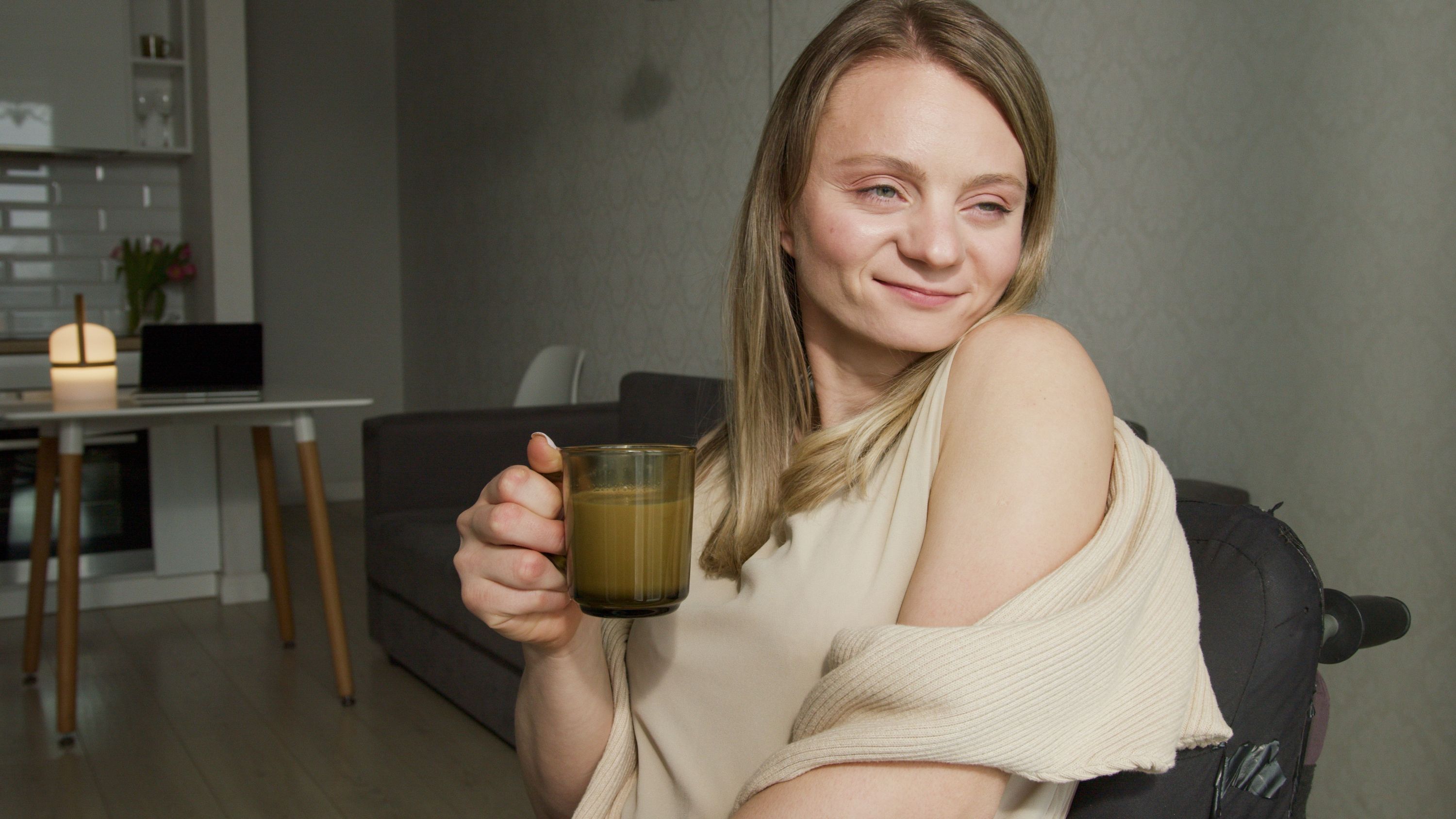 A woman with glasses pouring brewed coffee from a glass French press into a green mug inside a home kitchen.