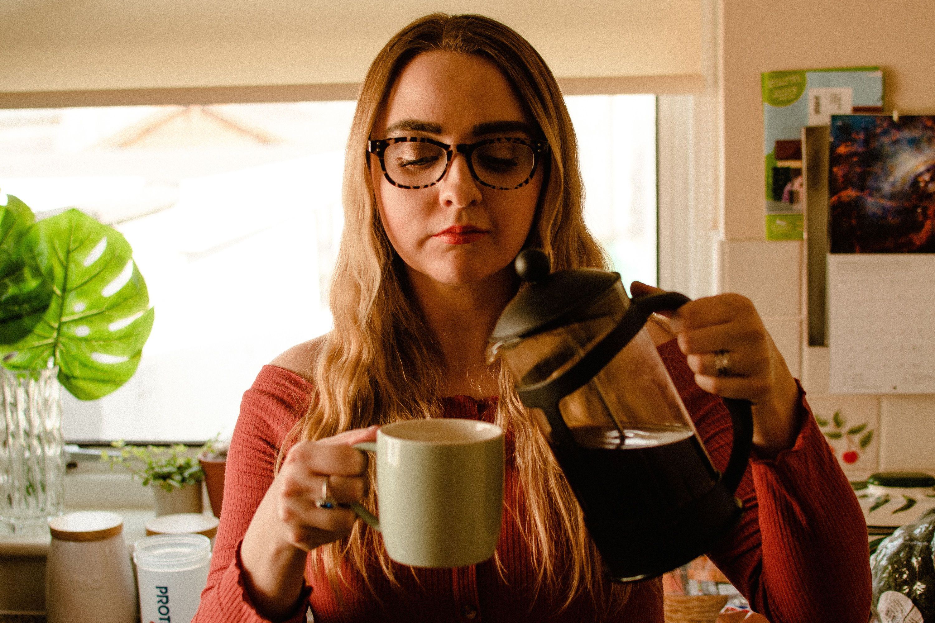 A woman with glasses pouring brewed coffee from a glass French press into a green mug inside a home kitchen.