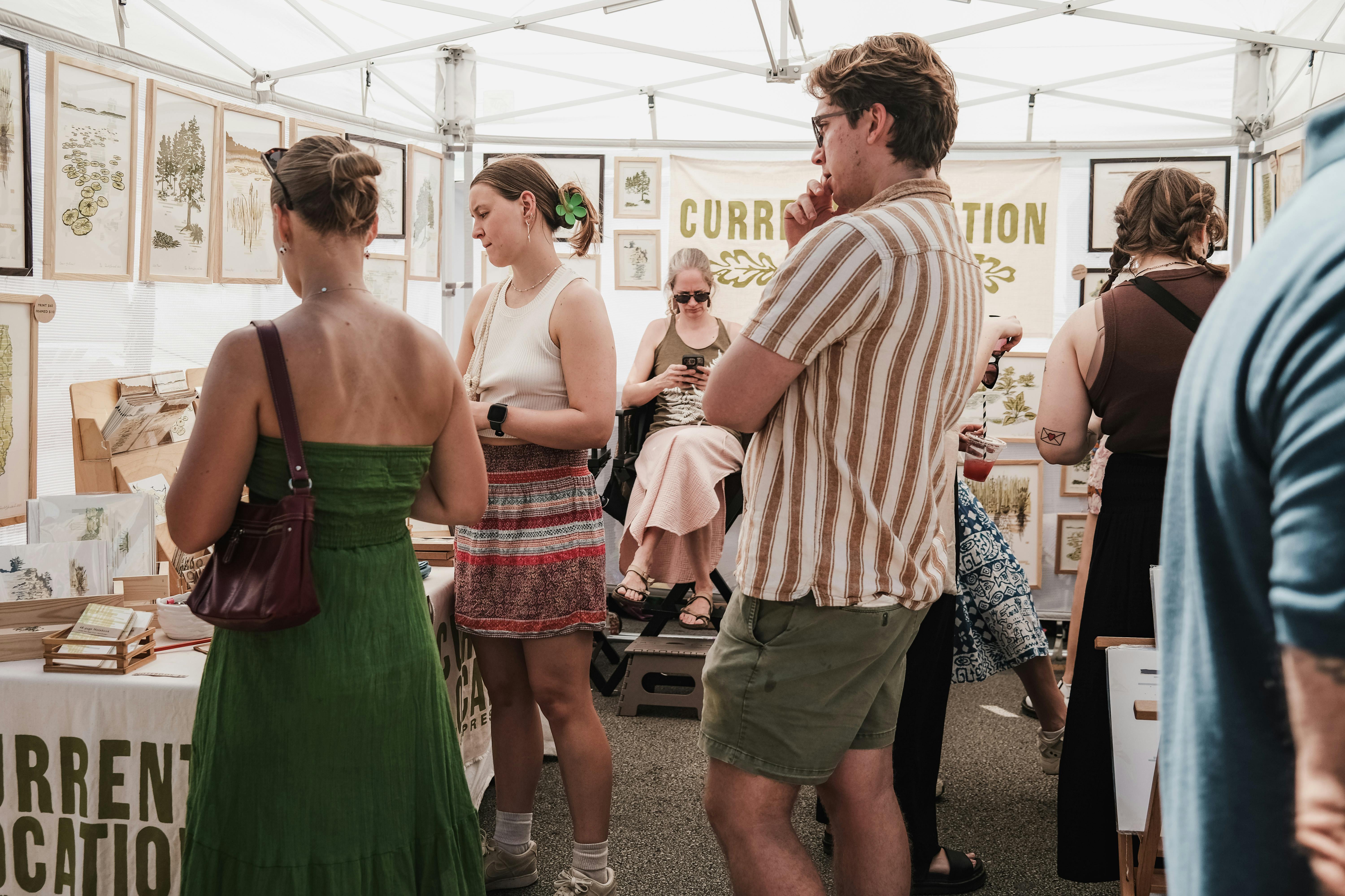 People wearing casual clothing standing inside a white vendor tent looking at framed artwork hanging on displays.