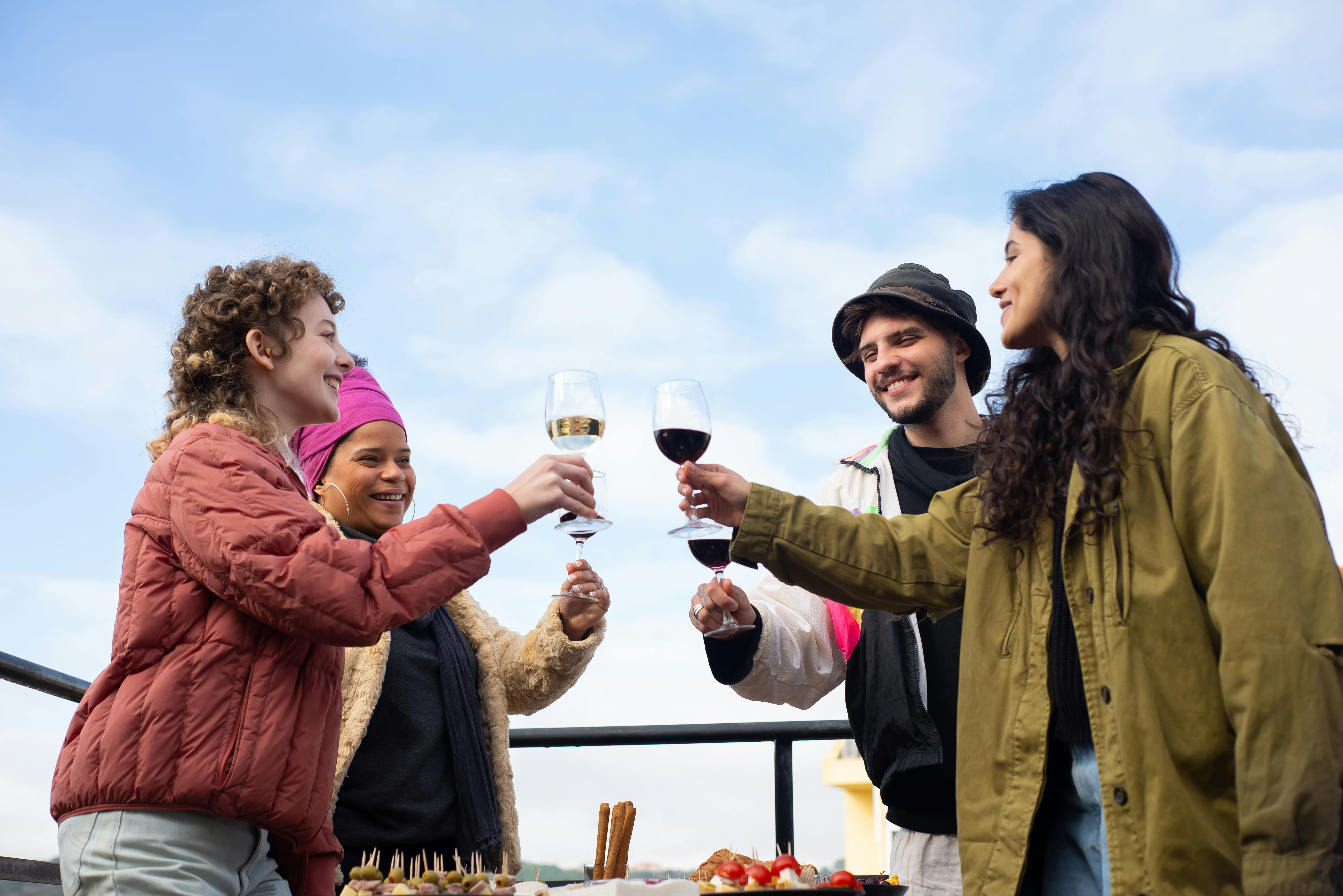 Smiling friends wearing casual clothing standing outdoors raising wine glasses against a bright cloudy sky.