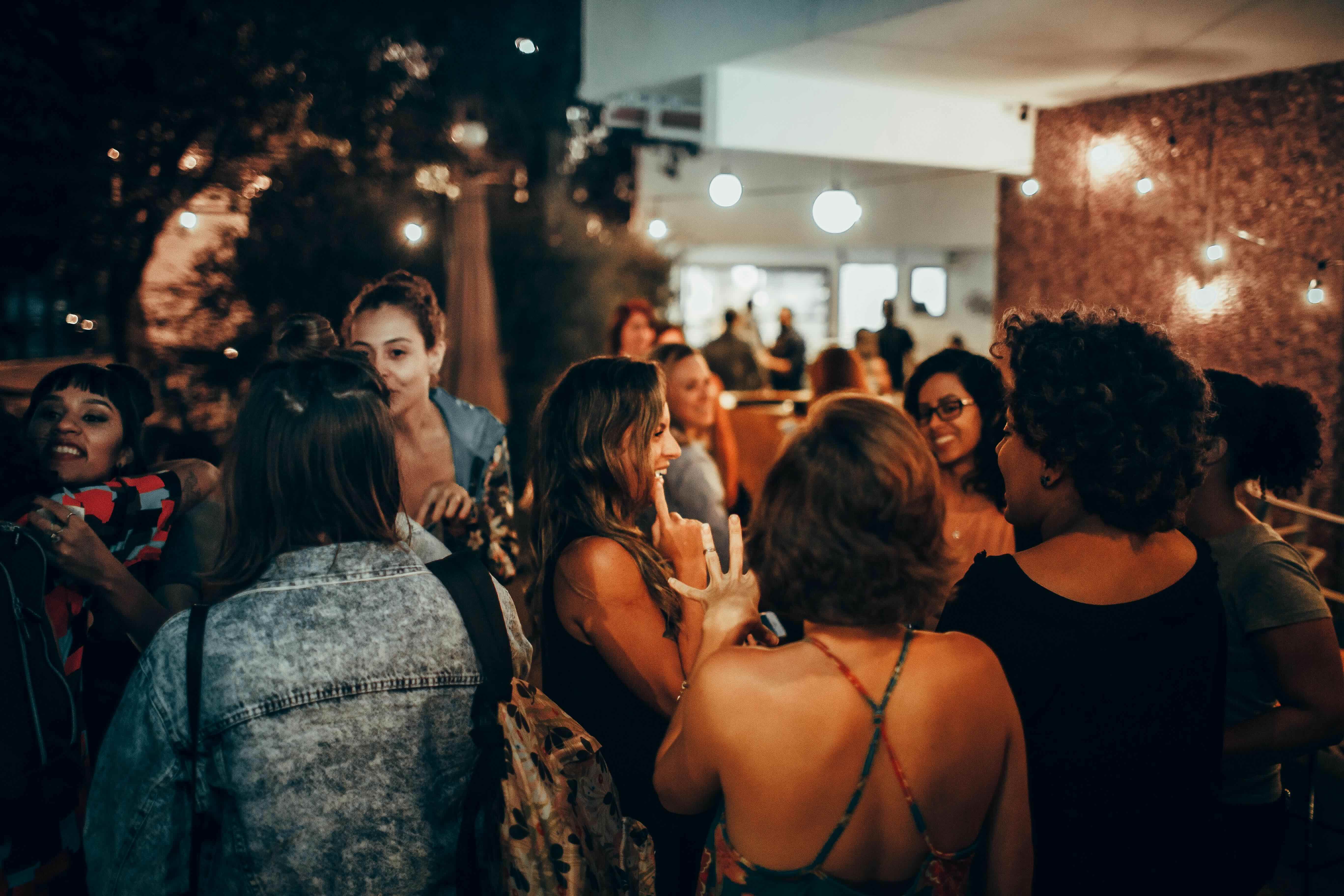 A large group of people standing and smiling while talking together outside a brick building under string lights.