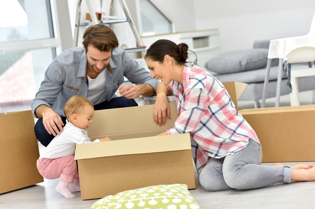 A smiling couple and their baby are on the floor opening moving boxes in their new rental home in Foley, Alabama.