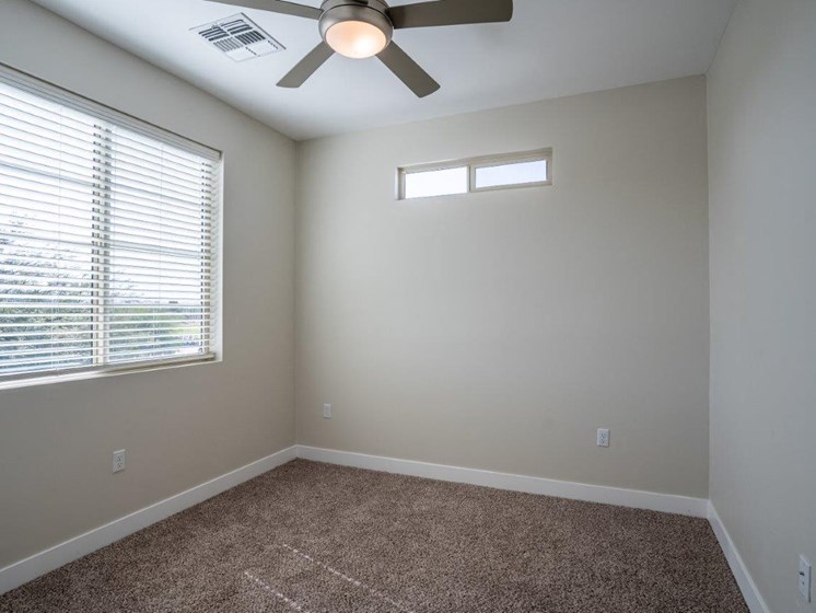 A carpeted bedroom has a window and ceiling fan in an apartment at Village Greens of Queen Creek
