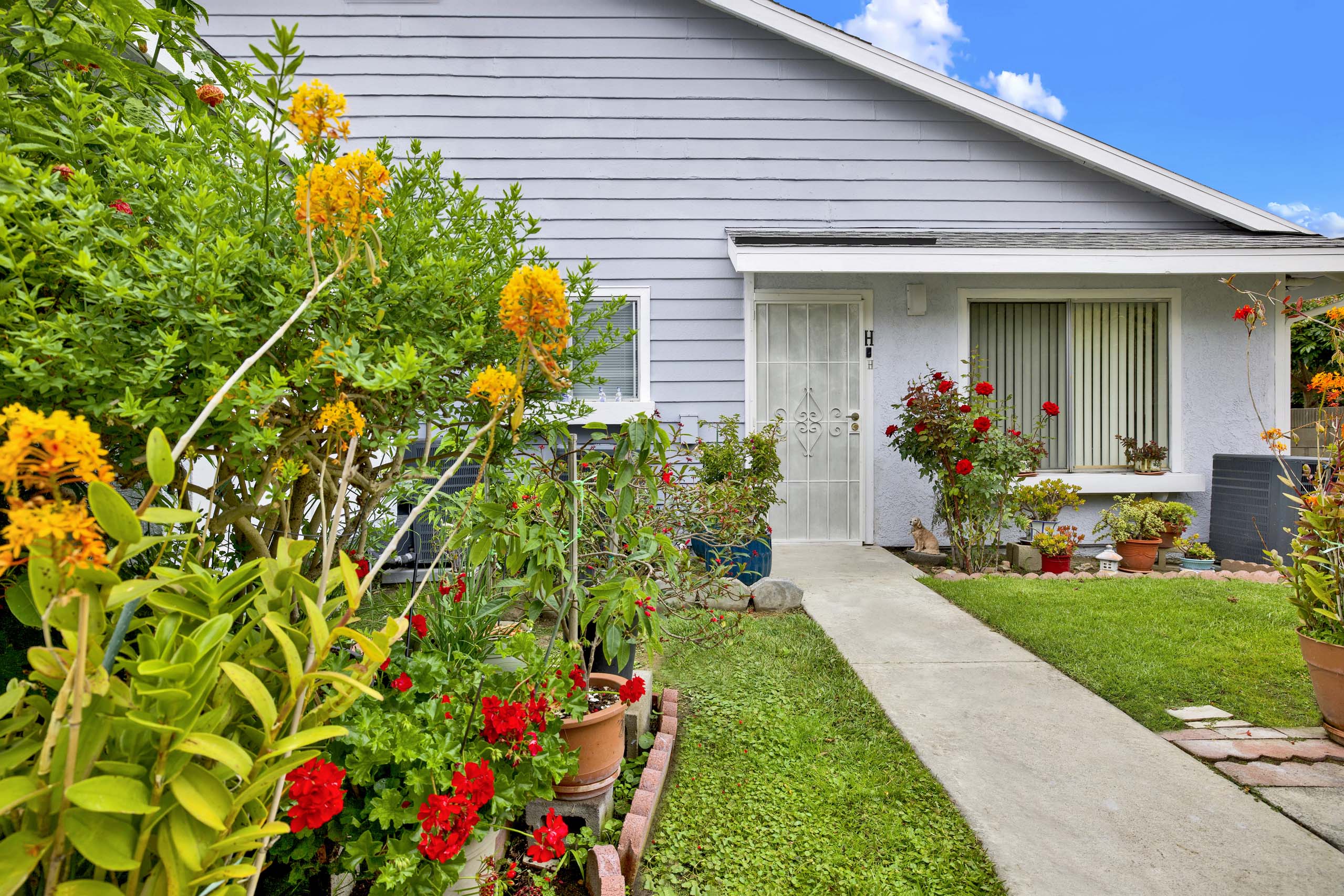 A house with a grey siding and a white door is surrounded by a variety of colorful flowers.