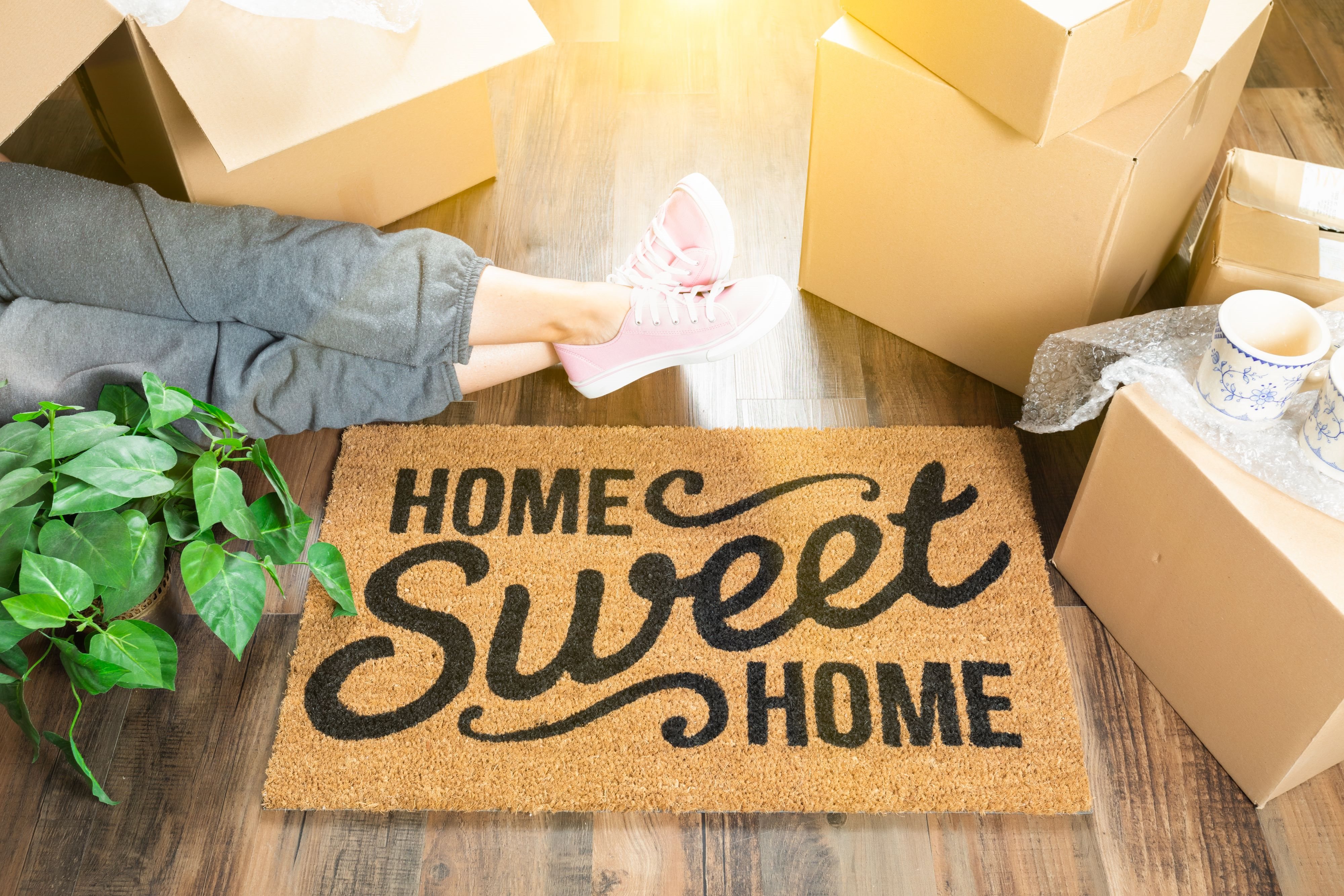 A person is unpacking boxes on a doormat that says "Home Sweet Home".