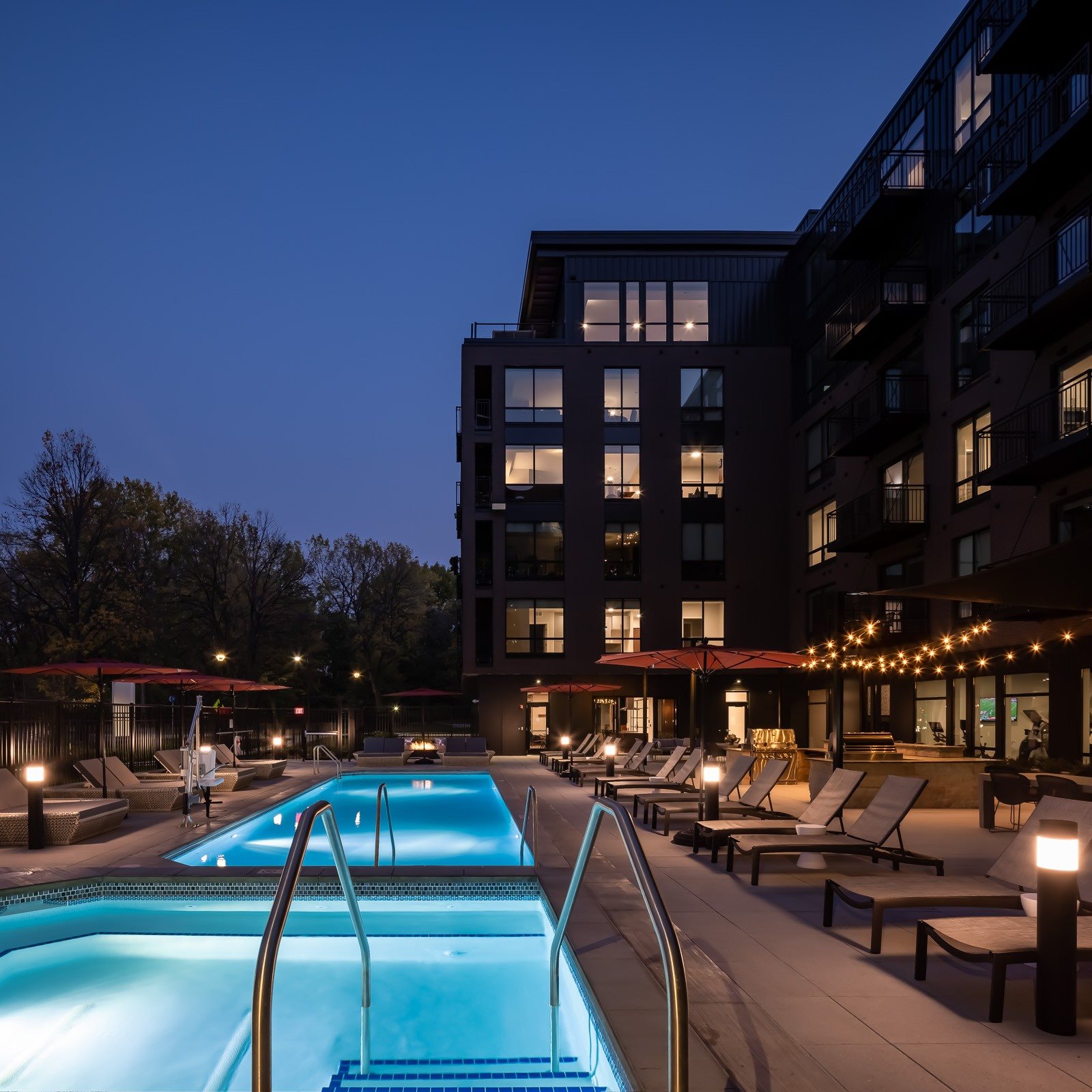 Dusk view of hot tub, pool, and pool seating on terrace with lit apartment windows