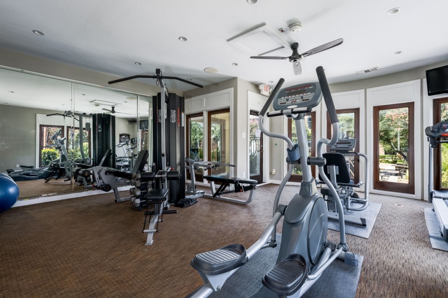 Spacious gym featuring mirrored wall, cable station, and cardio equipment by windows.