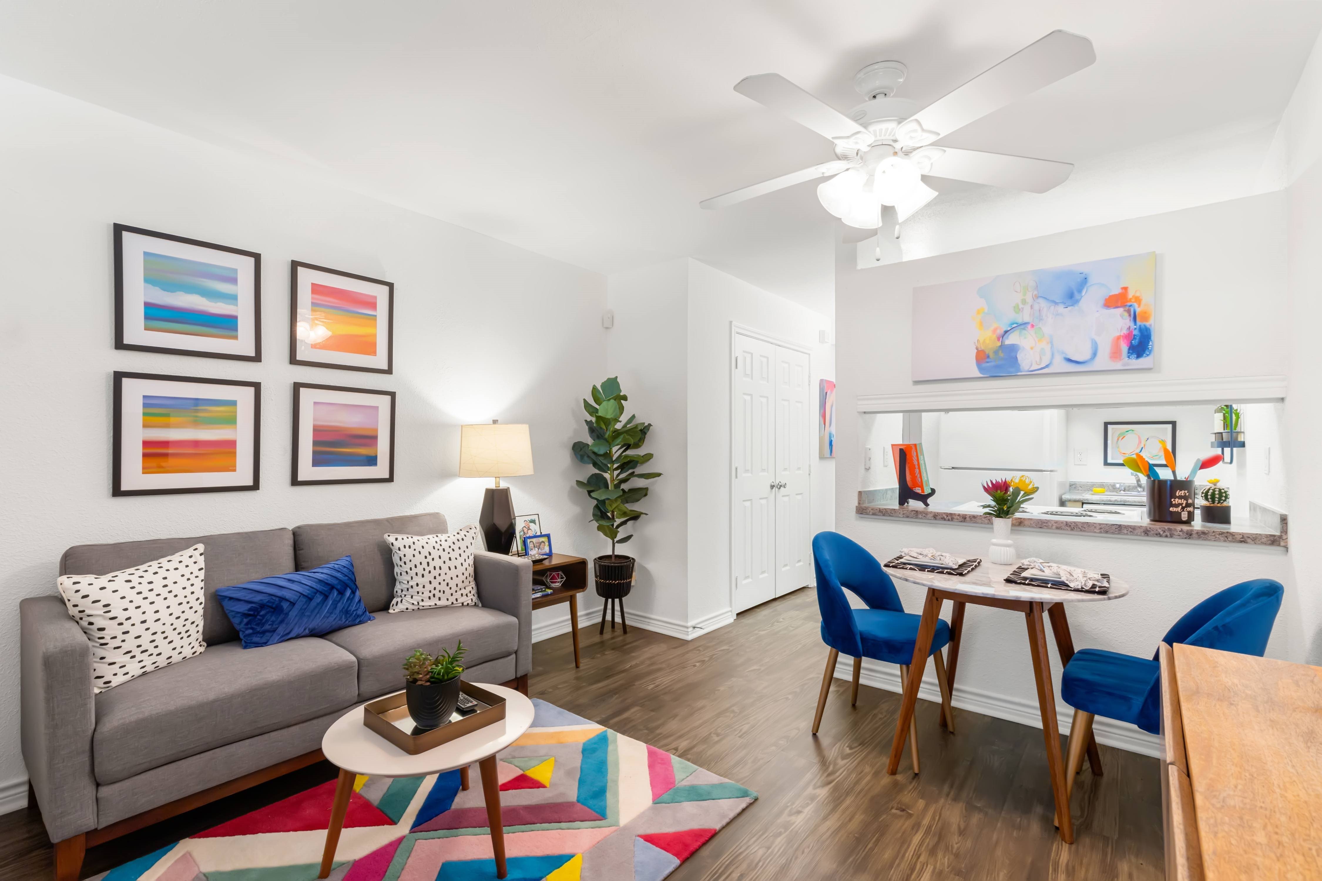 A living room with a grey couch, a coffee table, a dining table with chairs, a ceiling fan, and a painting on the wall.,A living room with a grey couch, a coffee table, a dining table with chairs, a ceiling fan, and a painting on the wall.