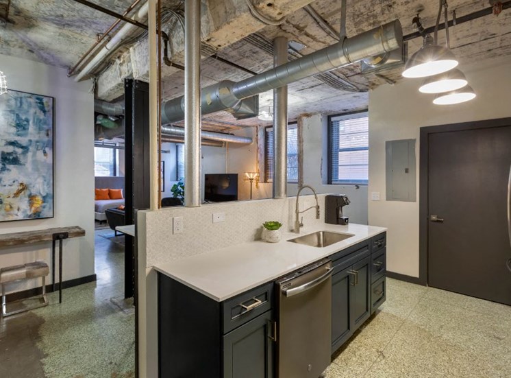 Industrial Style Apartment with Exposed Concrete and Ducts, Kitchen with White Counters, Blue Cabinets, Bar Style Table with Stools Under Art with Open Layout in the Background