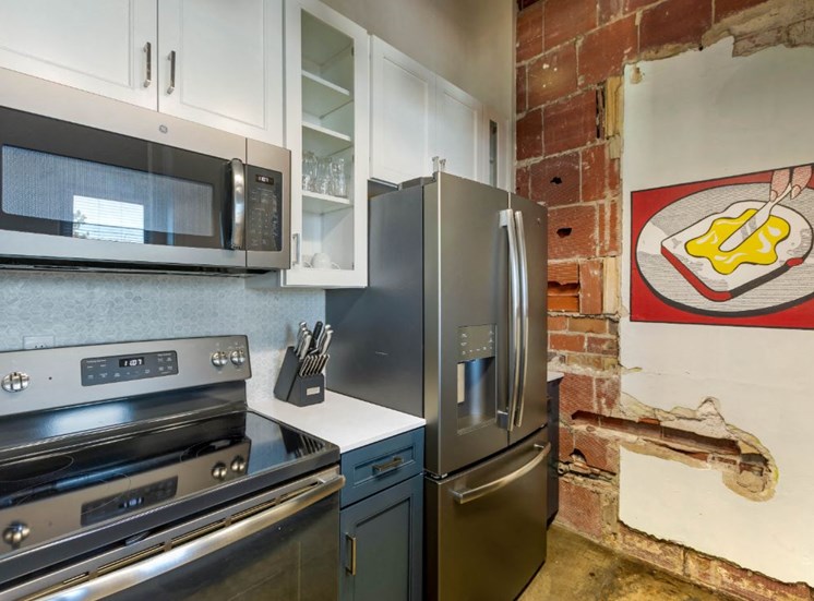 Industrial Style Apartment with Exposed Concrete and Ducts, Kitchen with Breakfast Bar, White Upper Cabinets and Dark Blue Lower Cabinets Around Stainless Steel Appliances Next to Exposed Brick and Art