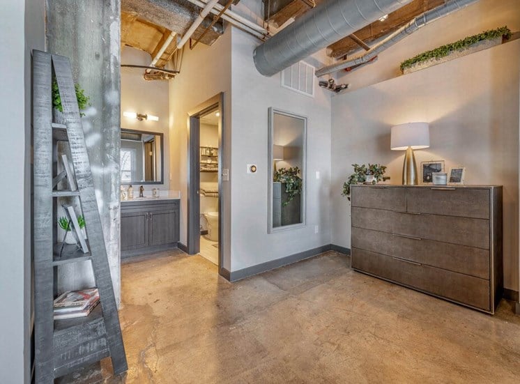 Industrial Style Apartment Bathroom with  Exposed Concrete, Decorative Shelf, Grey Wood Cabinets and White Cabinets Under Mirror Next to Bathroom Door and Full Length Mirror NExt to Dress with Plants and Decorations