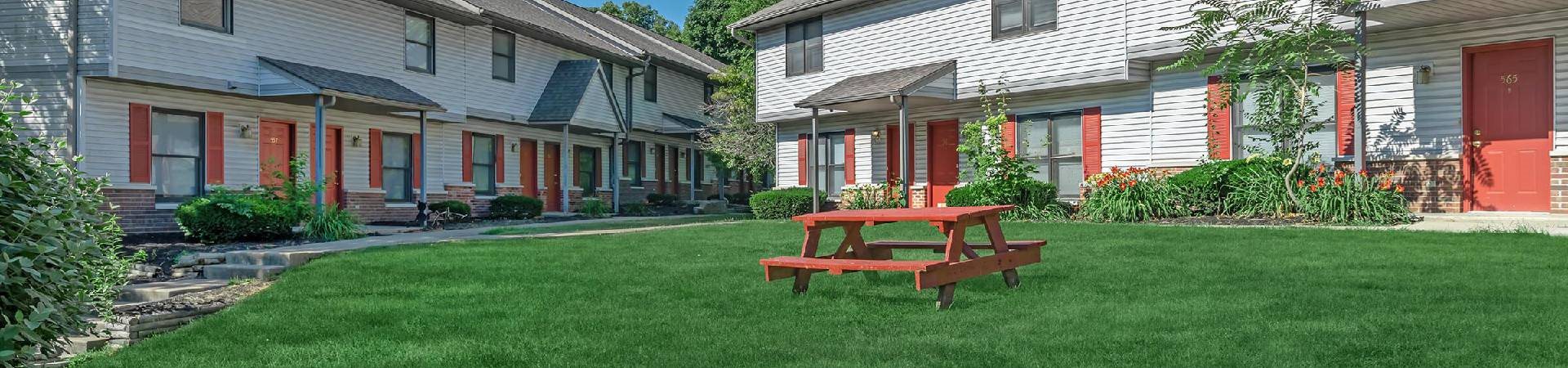 A red picnic table is in the middle of a grassy area in front of a white house.
