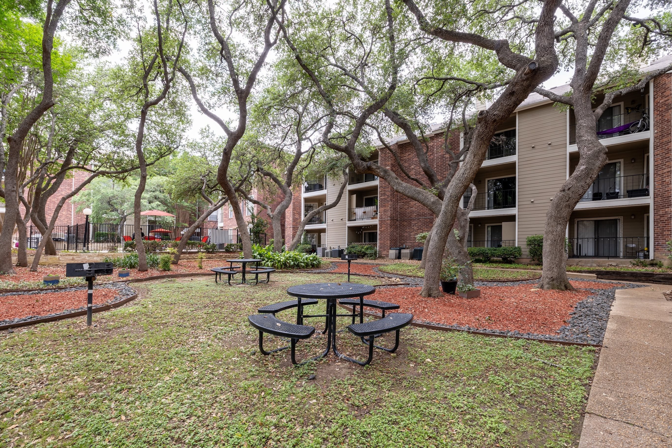 A park area with picnic tables and trees in front of apartment buildings.