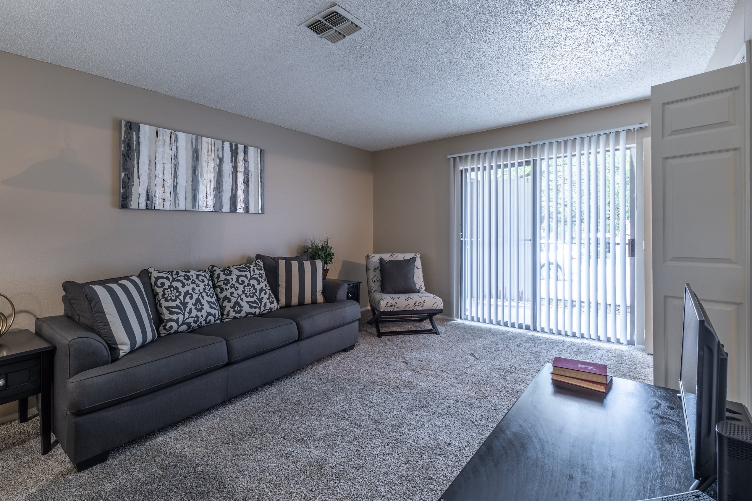 A living room with a grey couch, a small table, a television, and a window with blinds.