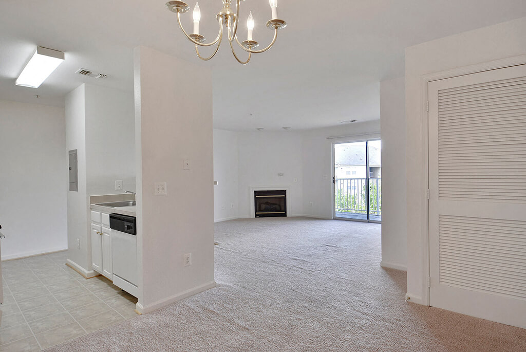 A large, empty living room with a chandelier and a kitchen area in the background.