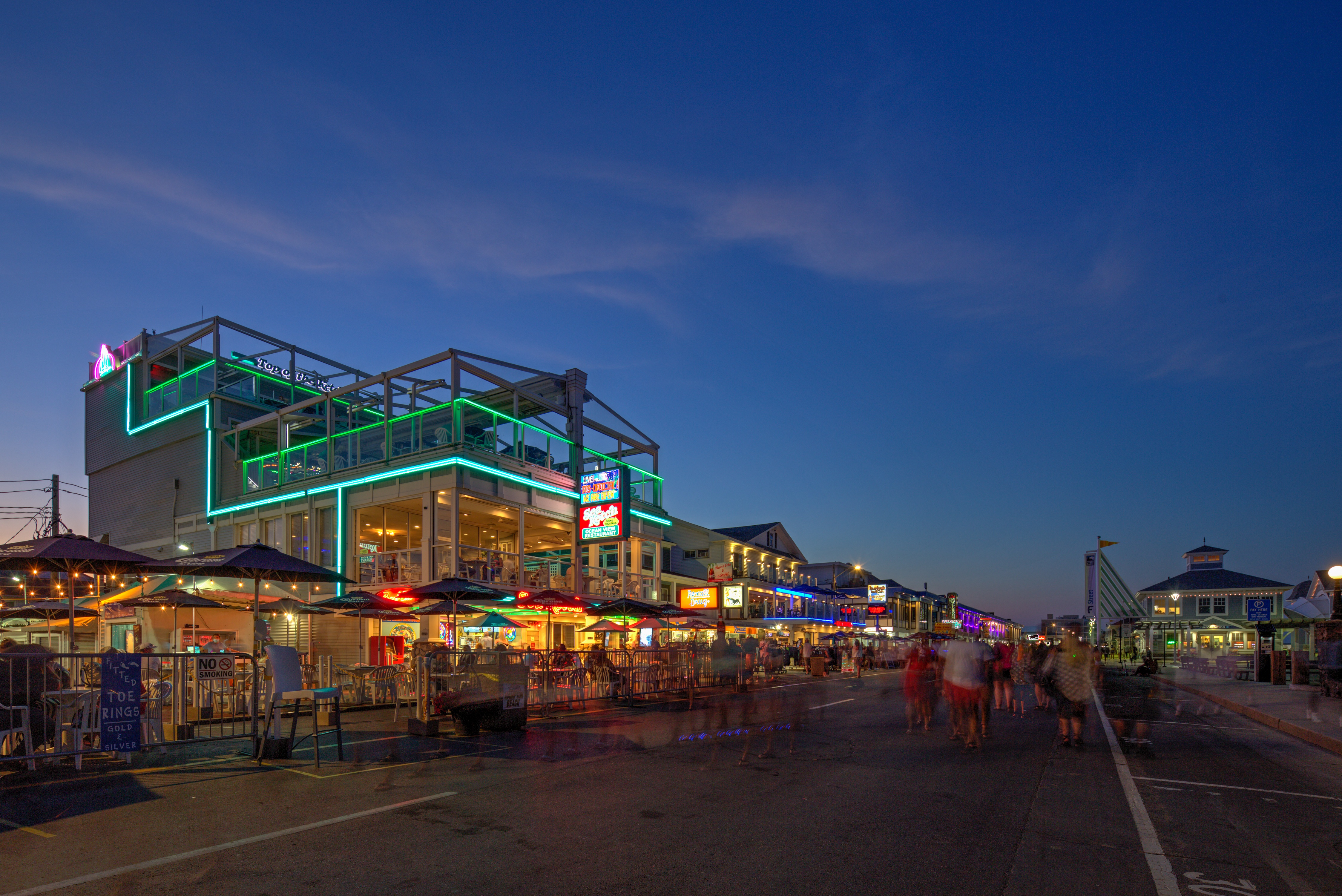 A group of people are walking on a street with a building with a green neon sign on it.