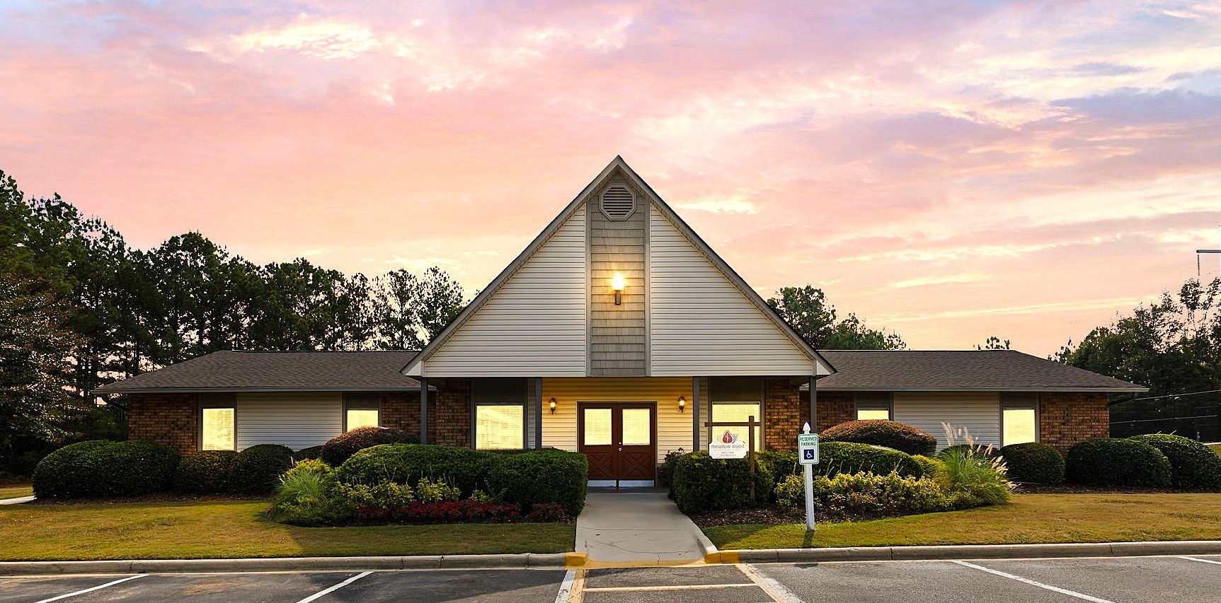 A church with a steeple and a cross on top is surrounded by bushes and has a parking lot in front.